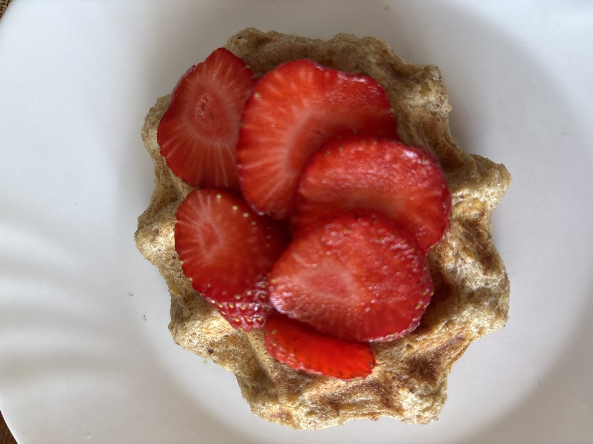 A close-up, overhead shot shows sliced strawberries atop a single, round waffle. The waffle is golden brown and textured with square indentations. The strawberries are bright red and sliced thinly, revealing their seeds. The waffle and strawberries are placed on a white plate. The background is out of focus and appears to be a white surface. The image was taken in Sochi, Russia.