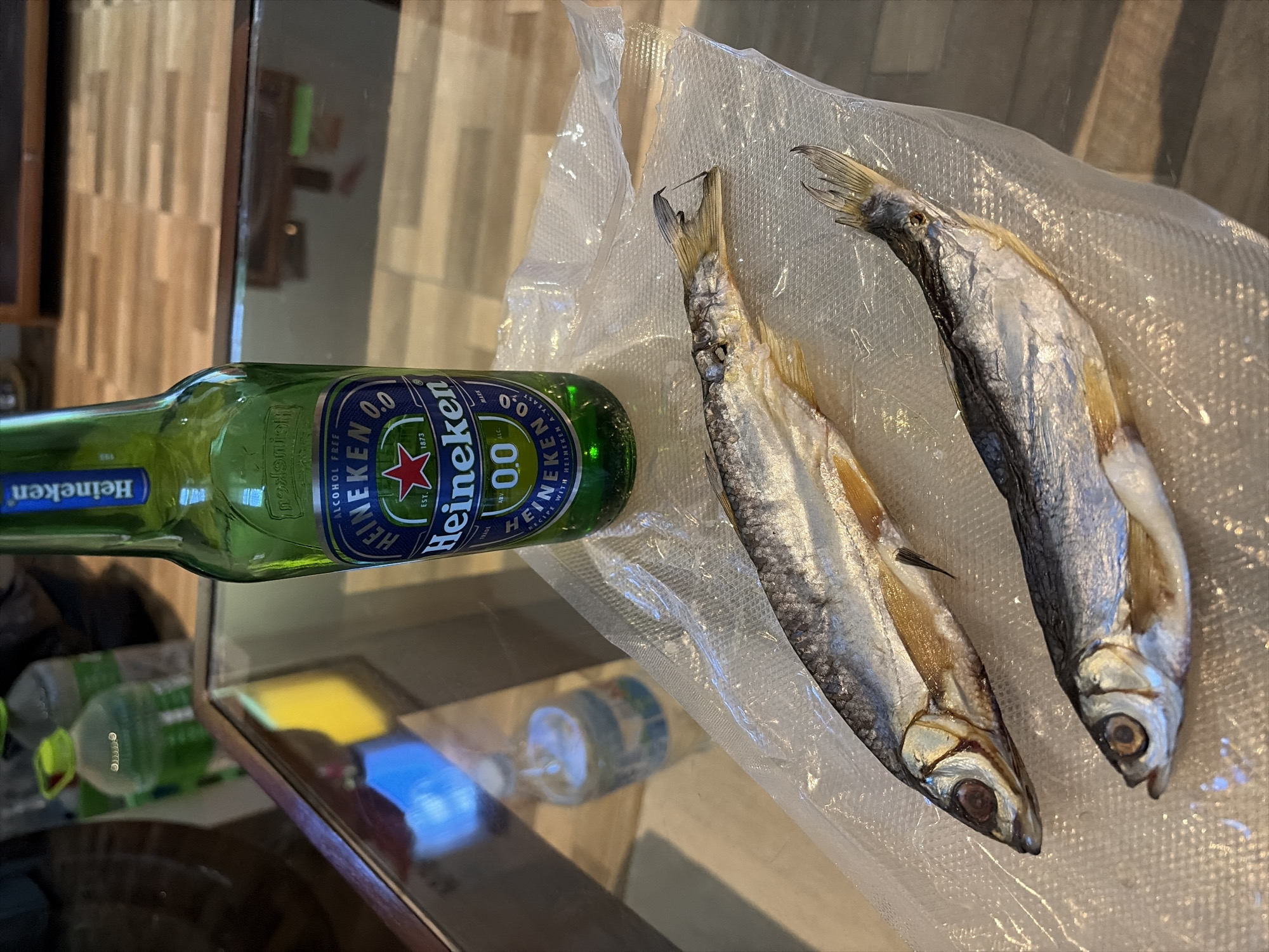 Two dried fish are laid out on a clear plastic sheet on a reflective glass table. Behind the fish, a green bottle of Heineken 0.0 beer stands upright. In the background, blurred bottles and a wooden floor are visible. The scene appears to be indoors, possibly in a residential setting within Baan Thong Po, Thailand, given the context provided.