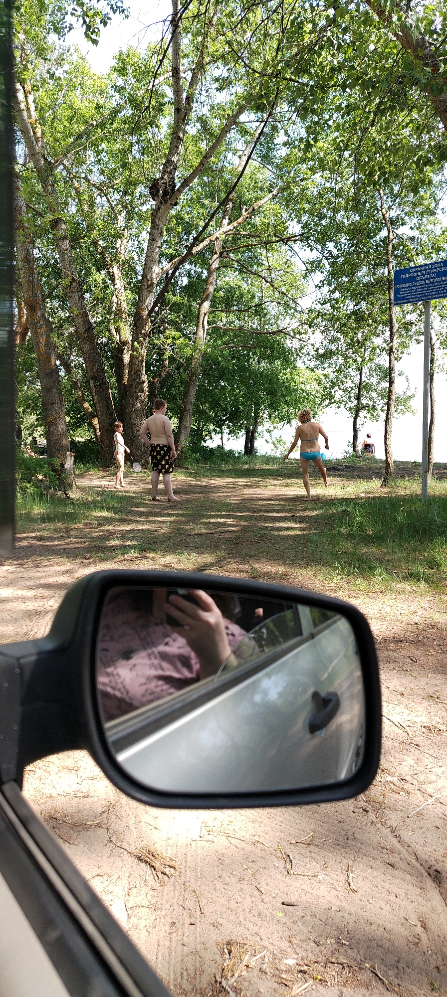 From the vantage point within a vehicle, the image captures a scene near a body of water in Noviy, Russia. Three individuals are visible near trees. One is a child with a slender build. To the right, a person with short hair and a blue swimsuit is running. Further to the left and slightly behind, a third individual in black and yellow swimming trunks stands still. There is a small sign by the water. The vehicle's side mirror reflects the person taking the photo.