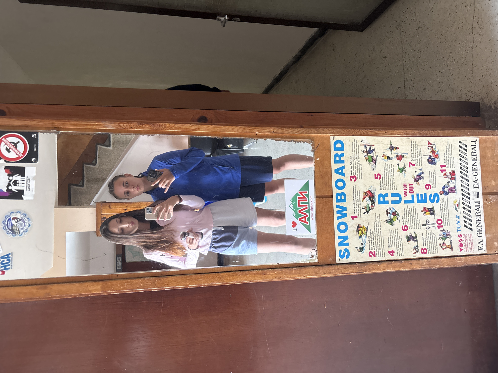 Two young women are taking a mirror selfie in an indoor hallway. The woman on the left is wearing a light pink jacket and grey and white shorts. She is holding a drink in her right hand and her left hand is on her hip. The woman on the right is wearing a blue jacket and dark shorts. She is making a hand gesture with her right hand. Behind them, a staircase is visible, along with various posters and stickers on the wall. One prominent poster is an "SNOWBOARD RULES" chart. The environment appears to be a public building, possibly a hostel or administrative office, given the functional aesthetic and the presence of multiple notices. The setting is identified as being in the city of Bistritsa, Bulgaria.