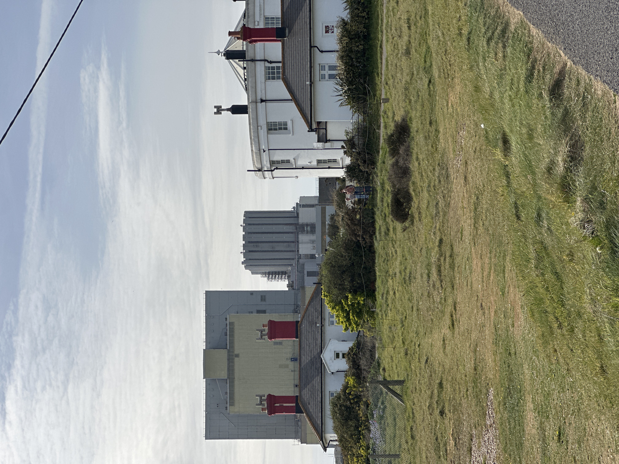 The image depicts a coastal scene, likely in the Folkestone and Hythe District, United Kingdom, given the distinct architecture and terrain. A white lighthouse with a red top stands prominently, positioned near grassy terrain that slopes down to a road or pathway. Further back are large, industrial buildings, suggesting a mix of residential and industrial areas. Two individuals are visible near a fence line and some greenery, seemingly engaged in a conversation. The sky is overcast with clouds. An electrical cable is visible in the top left of the frame.