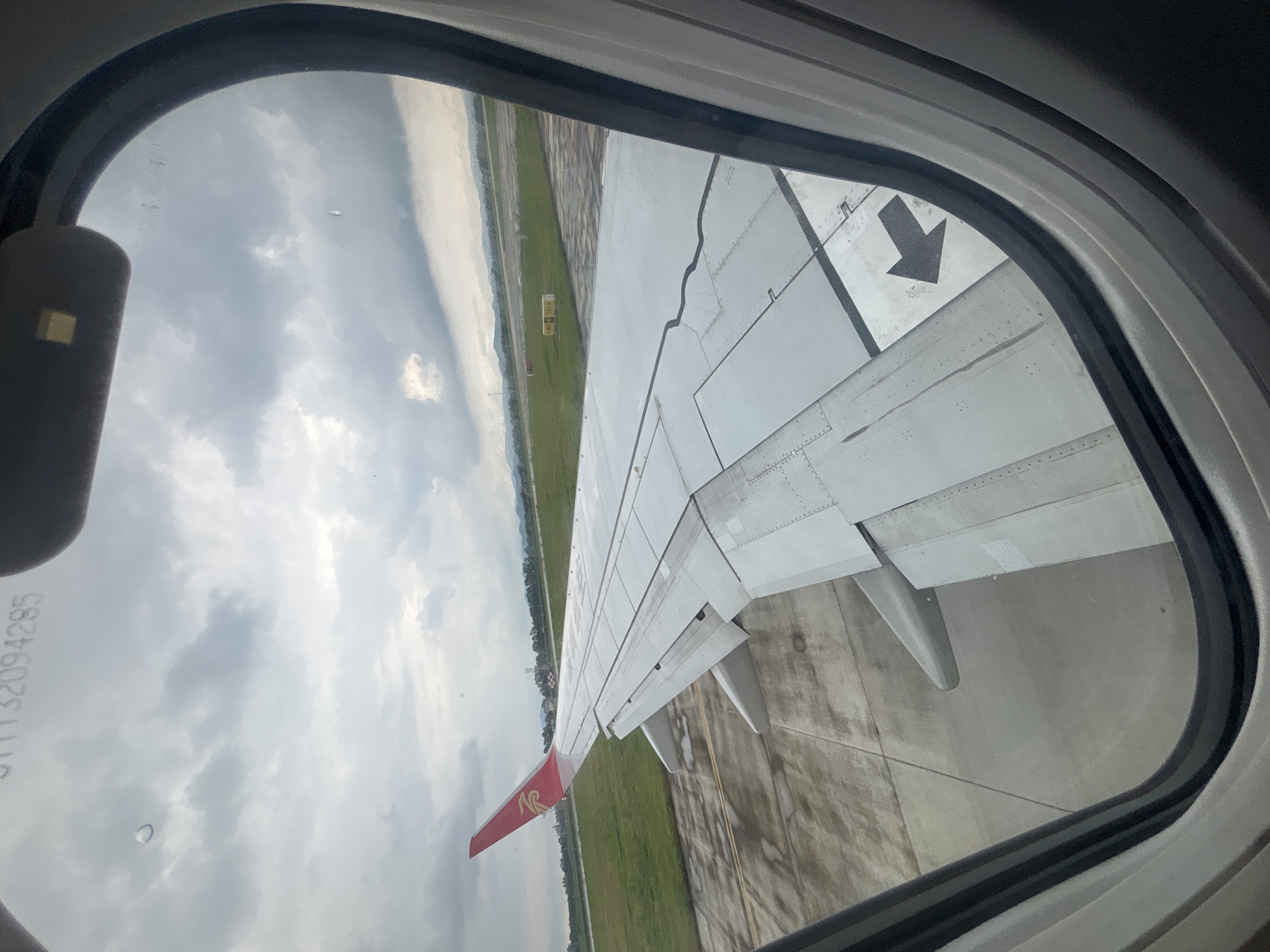 The image is taken from an airplane window, looking out at a cloudy sky and an airport tarmac. An airplane wing, with its red and white tailfin, dominates the foreground. The wing is metallic and shows signs of wear. A black arrow is painted on the wing's surface, pointing to the left. In the background, there is a green field and some distant mountains. The tarmac is wet, suggesting recent rain. The view is framed by the oval shape of the airplane window. There are no people visible in the image. The overall scene depicts a moment before takeoff or after landing at an airport.