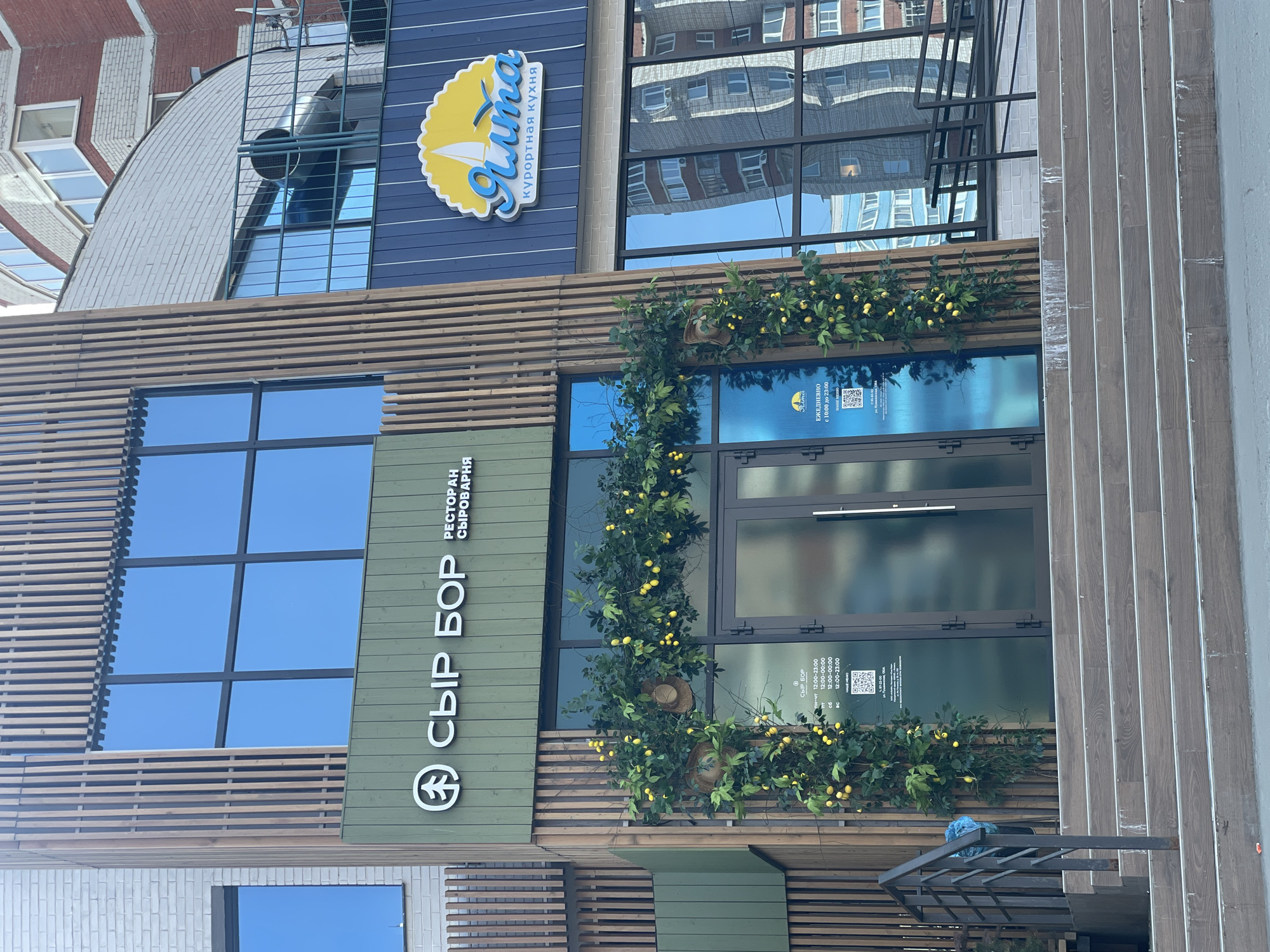 The image depicts the exterior of a restaurant called "Cheese Bor" (СЫР БОР) in Izhevsk, Russia. It is a cheese-making restaurant ("RESTORAN SYROVARNYA"). The building has a modern design with large windows and horizontal wooden slats on the facade. The entrance is adorned with greenery and small yellow decorations. Above the restaurant is another sign for "Yalta" (Ялта), a resort kitchen, with a logo of a yellow sun with a sailboat. The upper part of the building is blue, complementing the restaurant below. The sky is reflected in the windows, and part of another building is reflected in the upper windows. There is a metal railing on the right side of the building.