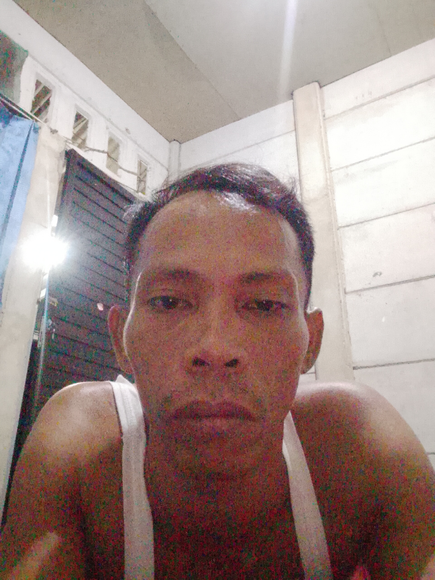A man is centered in the frame, wearing a white sleeveless shirt. He is indoors, looking directly at the camera. Behind him is a door, with a bright light source above it. The walls appear to be constructed of concrete blocks. The location is likely Bukit Ketok, Indonesia. The image is grainy.