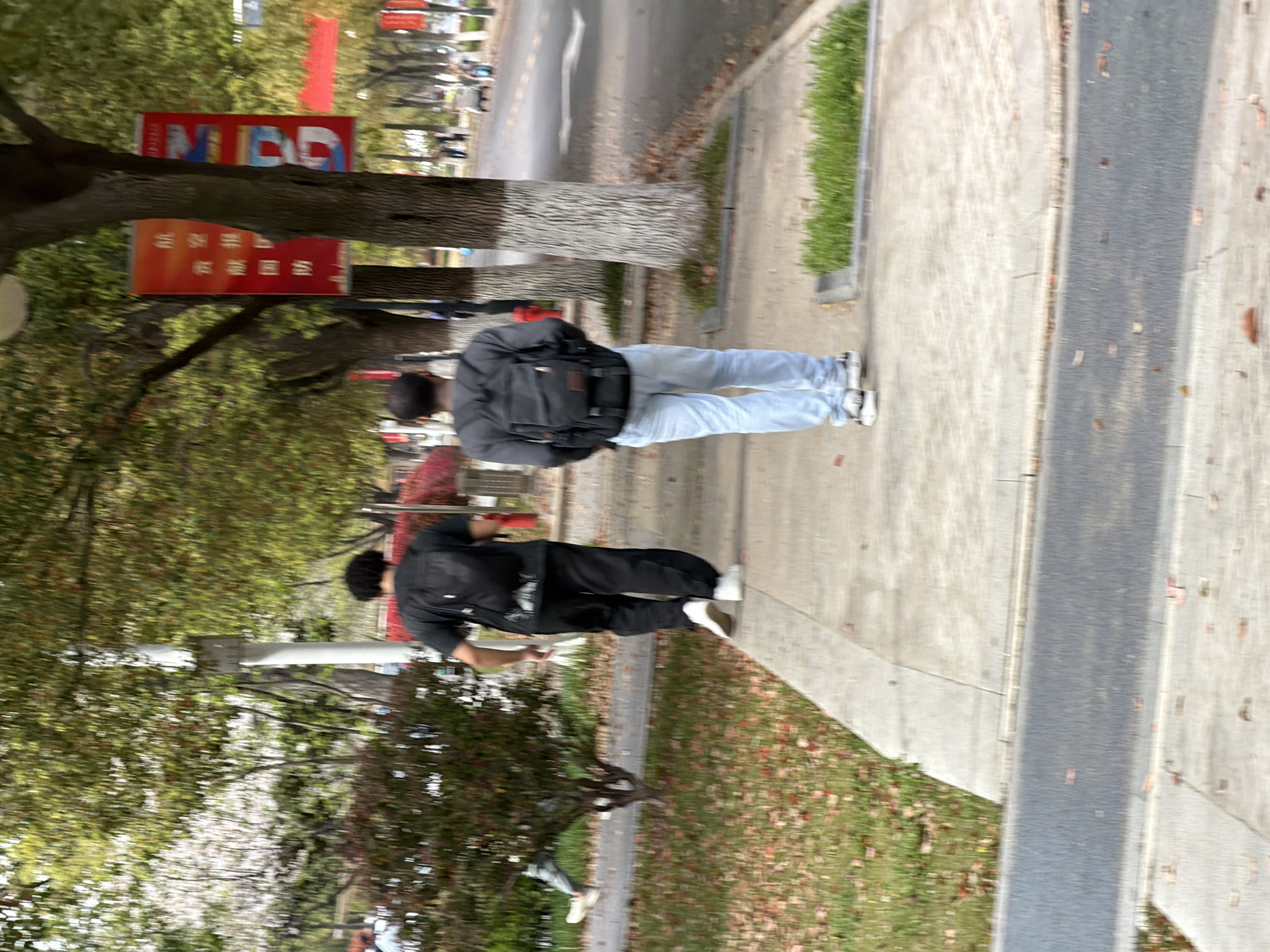 The image shows two individuals walking along a sidewalk in what appears to be a park or campus setting in Jiangning District, China. The sidewalk runs alongside a street and is bordered by trees and grass. The first individual is wearing a dark jacket and black pants. The second individual is wearing a dark jacket and light-colored jeans and carrying a backpack. Signs in Chinese are visible on nearby trees.