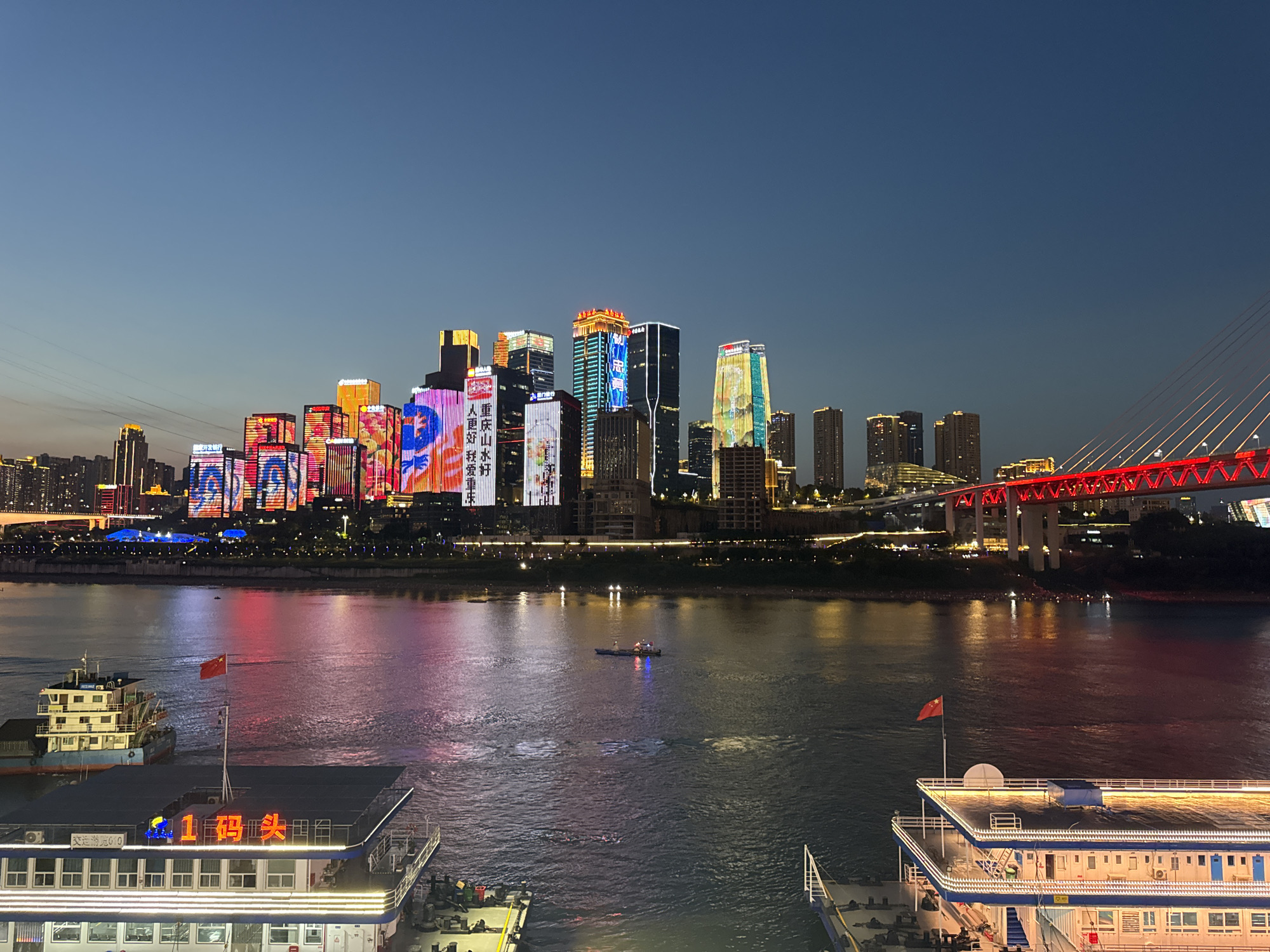 The image depicts a night view of a cityscape in Yuzhong District, China, situated along a wide river. Dominating the skyline are numerous illuminated skyscrapers, their facades displaying vibrant, colorful lights and Chinese characters. A prominent red suspension bridge with illuminated cables spans across the river on the right side of the frame. In the foreground, the tops of two large passenger ships are visible, docked or moored on the water. One ship, on the left, has a Chinese flag flying from its mast. The river's surface reflects the bright city lights, creating a shimmering effect. The sky is a deep, dusky blue, indicating either dusk or dawn. The overall scene is a bustling urban environment at night, characterized by impressive architecture and transportation infrastructure.