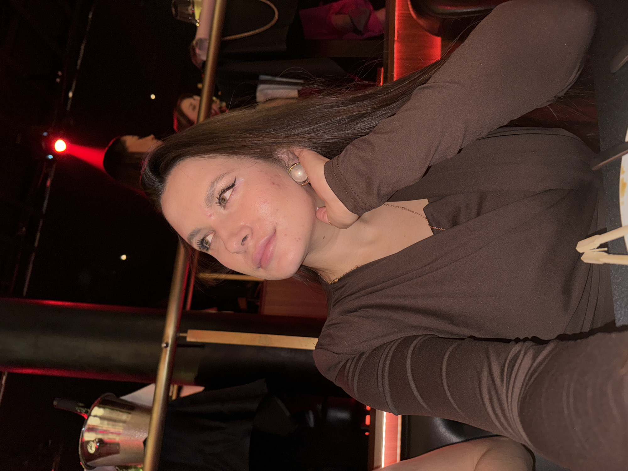 A woman is seated at a table in a dimly lit venue. Her brown hair falls over her shoulder, and she is wearing a brown, long-sleeved top and a gold necklace. She is holding her left ear. The lighting suggests a nightlife setting, possibly a bar or restaurant. An ice bucket is visible on the left side of the frame. The location is suspected to be in the city of Belgrade, Serbia.