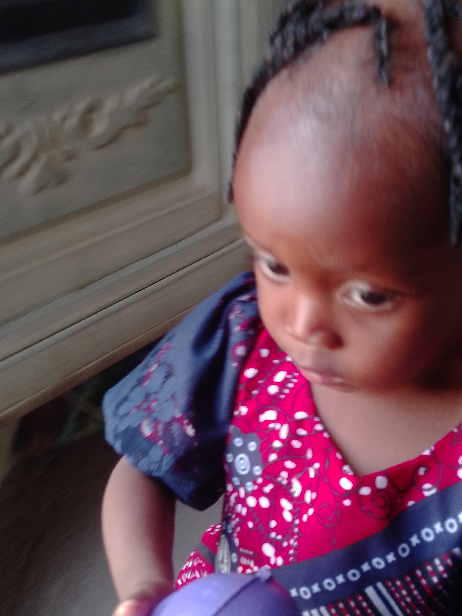 A young child with braided hair is looking down at a purple object they are holding. The child is wearing a red and white patterned shirt with blue sleeves that have a lighter blue floral design. A piece of furniture, possibly a cabinet or dresser, is visible in the background. The location is unknown.