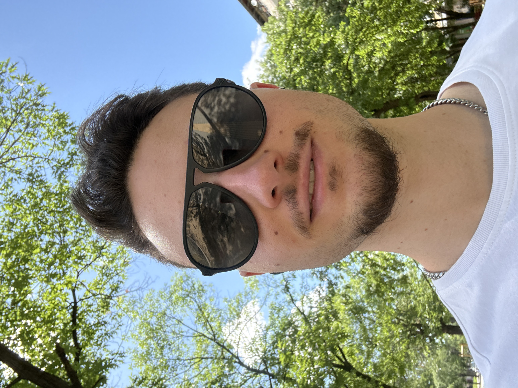 A man with dark hair and a beard is wearing sunglasses and a white t-shirt. He is outside, under green trees and a blue sky. He is wearing a silver chain necklace. The location is Sofia, Bulgaria.