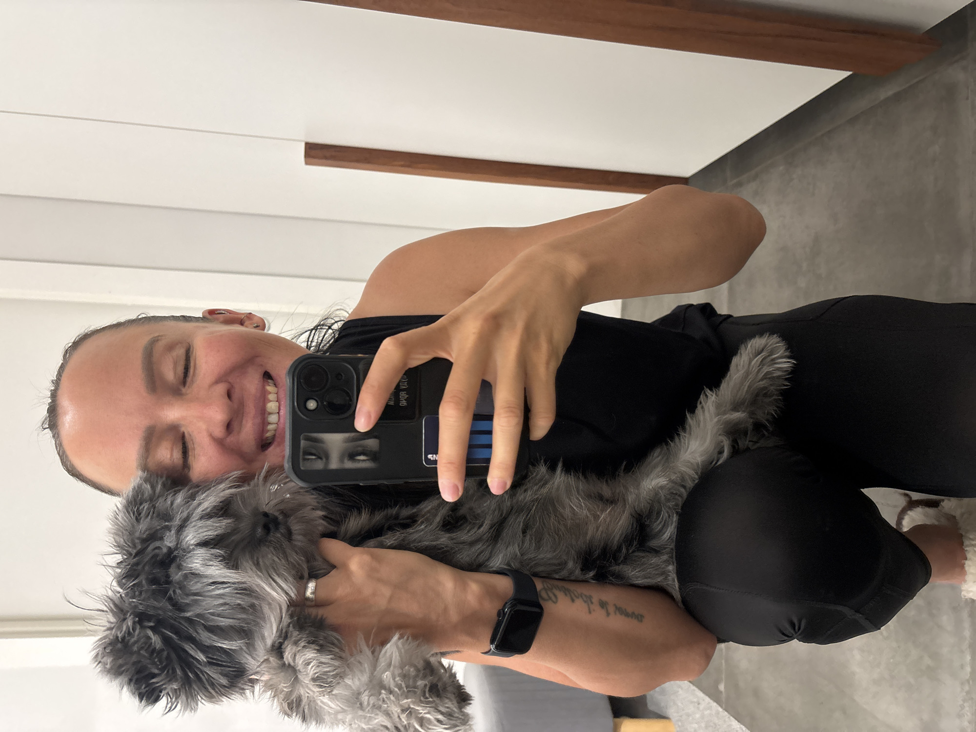 A woman is taking a selfie with a gray, long-haired dog. The woman is fair-skinned with brown hair pulled back, wearing a black tank top and black leggings. She is holding a black iPhone with a graphic case. She is smiling. She wears a watch on her left wrist. The dog is gray and furry, nestled in her arms.
The background consists of a white wall with a brown wood accent, a gray wall, and a gray floor. The location is Chaikovsky, Russia.