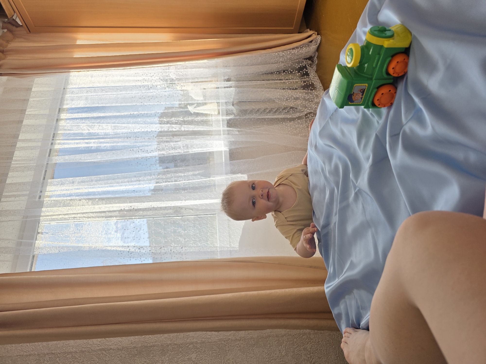 A baby is looking up from behind a blue bedsheet. A toy train is on the bed near the baby. A window with tan and white curtains is in the background. The location is Chaikovsky, Russia. A leg and foot are partially visible in the lower right.