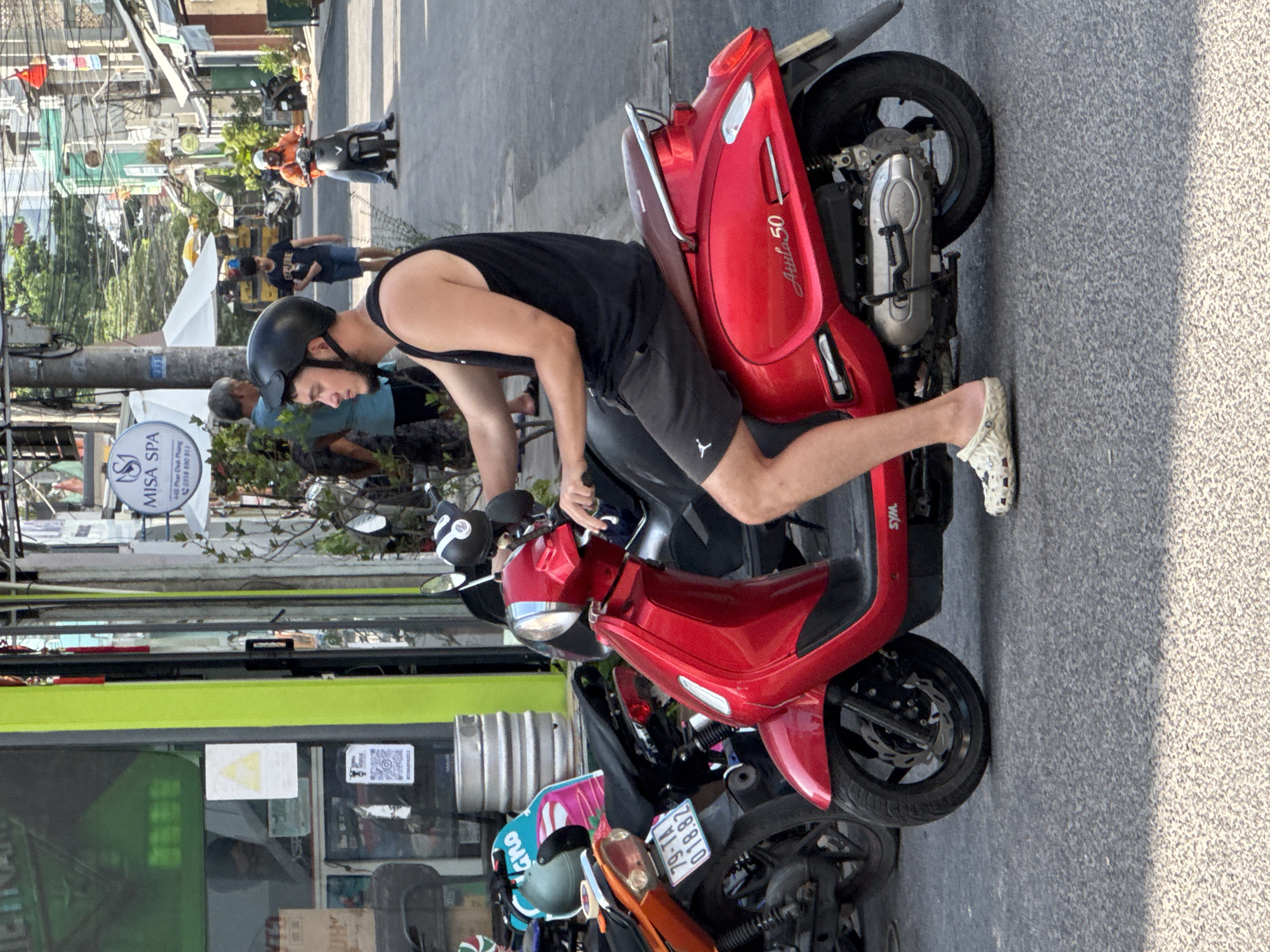 This is a medium-shot, eye-level, static street scene captured on a sunny day in Nha Trang, Vietnam, likely on a commercial street given the shopfronts and parked motorcycles. The primary subject is a young Caucasian male wearing a black helmet, a black tank top, and grey athletic shorts, seated on a bright red SYM Attila 50 scooter. He is looking slightly to his left. The scooter is parked or moving slowly on an asphalt road, with its front wheel visible and a portion of the rear wheel partially obscured. To the left of the red scooter, another scooter, orange and black, is parked with its license plate showing "79-TA 018.82". Further left, a shopfront with a bright green trim and a glass door displays various items inside, including stacked barrels. In the background, other individuals are present, some walking and others on motorcycles. A white canopy tent is visible further down the street, suggesting a commercial area. The architecture in the background indicates a dense urban environment with multi-story buildings. The overall scene is a typical snapshot of daily life in a Vietnamese city, characterized by the prevalence of scooters as a mode of transportation.