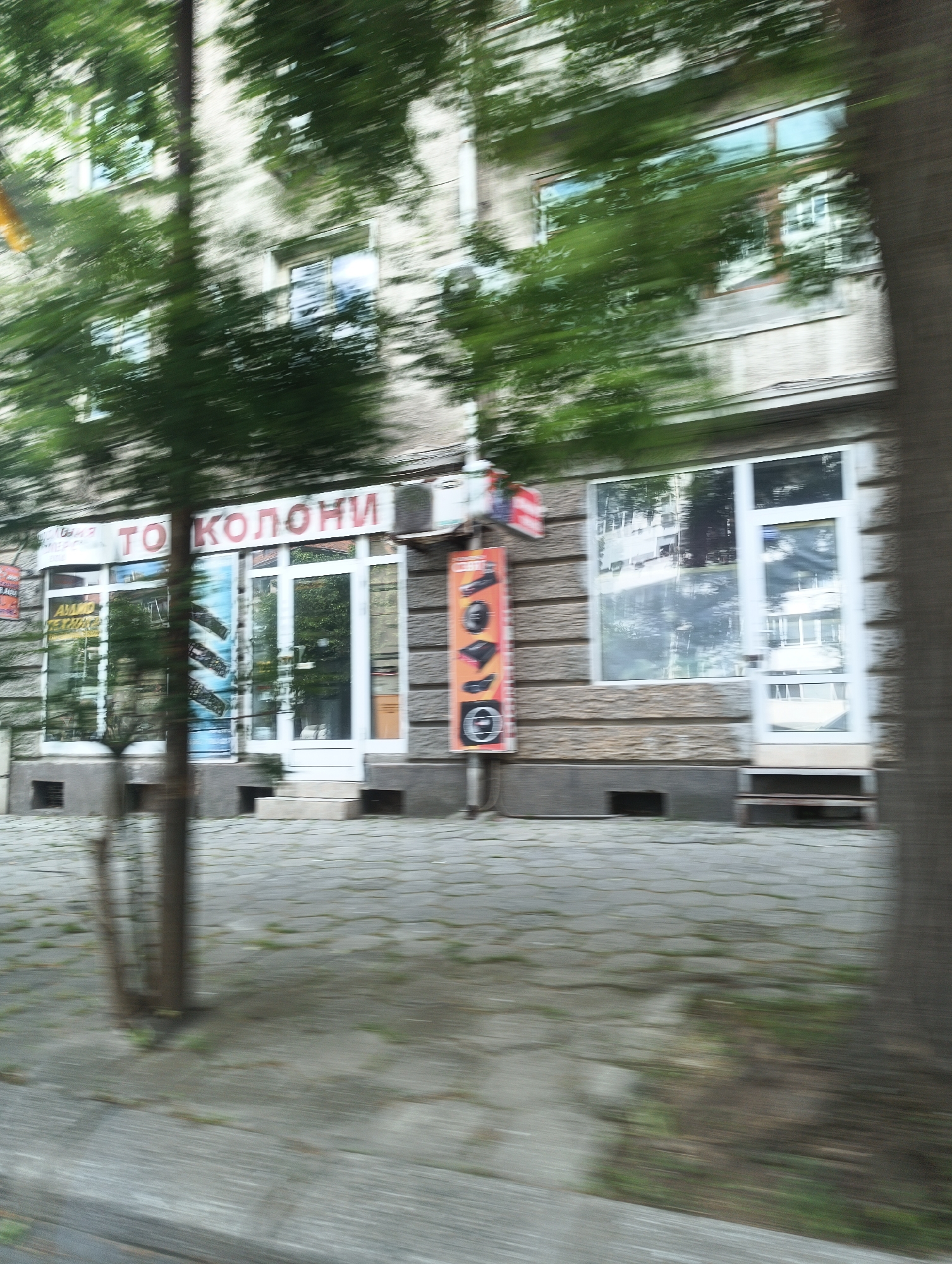 The image depicts a blurred street scene, likely taken from a moving vehicle. Dominating the frame is the facade of a building housing a store, identified by the signage above the entrance that reads "ТО КОЛОНИ" in Cyrillic script. This store likely sells car parts. The facade is textured with horizontal grooves, with windows and a white-framed door present. Adjacent to the store, there is a vertical orange sign showing images of car parts. The ground in front of the building is paved with hexagonal concrete blocks, displaying patches of greenery. Trees and foliage partially obscure the view of the building, adding to the sense of motion and blur. The location is likely Burgas, Bulgaria.