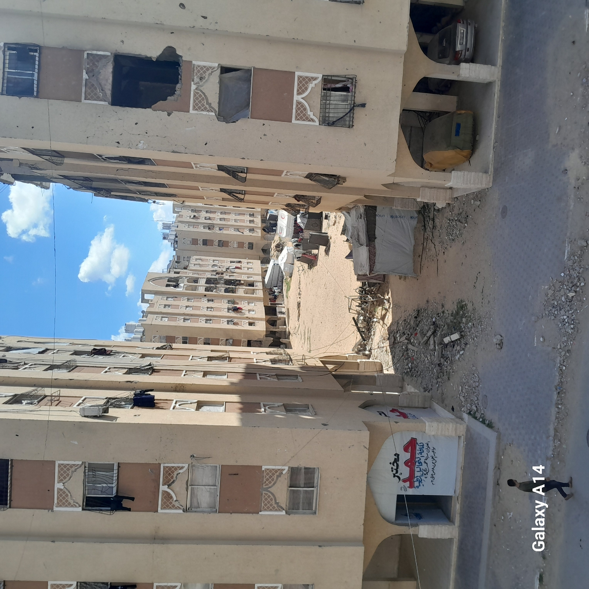 The image depicts a narrow street or alleyway flanked by multi-story residential buildings, captured during bright daylight under a blue sky with scattered white clouds. The buildings appear aged and show visible signs of damage, including large holes, broken windows, exposed rebar, and cracks in the plaster. Some windows are boarded up, while others have protective bars or clothes hanging out to dry. The architecture features light-colored facades (beige/sand) with some decorative tile patterns around windows.
In the open space between the buildings, several makeshift tents are visible, some white and one with "unicef" written on it, suggesting a humanitarian presence or displacement. The ground is a mix of sandy dirt and paved areas, littered with rubble and debris. A silver car is partially visible parked under an archway at the base of one building, and a large green and yellow bag sits nearby.
In the foreground, on the right side of the paved area, a lone person is seen walking away from the camera. On the ground floor of the building in the foreground, a large banner with Arabic text is displayed. The text translates to "Mukhtabar (Laboratory) Hamad," "All Medical Analyses," "Address: Bara City 25," and "Working hours from 8 AM to 2 PM," indicating a medical clinic or laboratory. The overall scene suggests a challenging living environment, possibly in an area affected by conflict or severe economic hardship. A "Galaxy A14" watermark is present in the lower right corner.