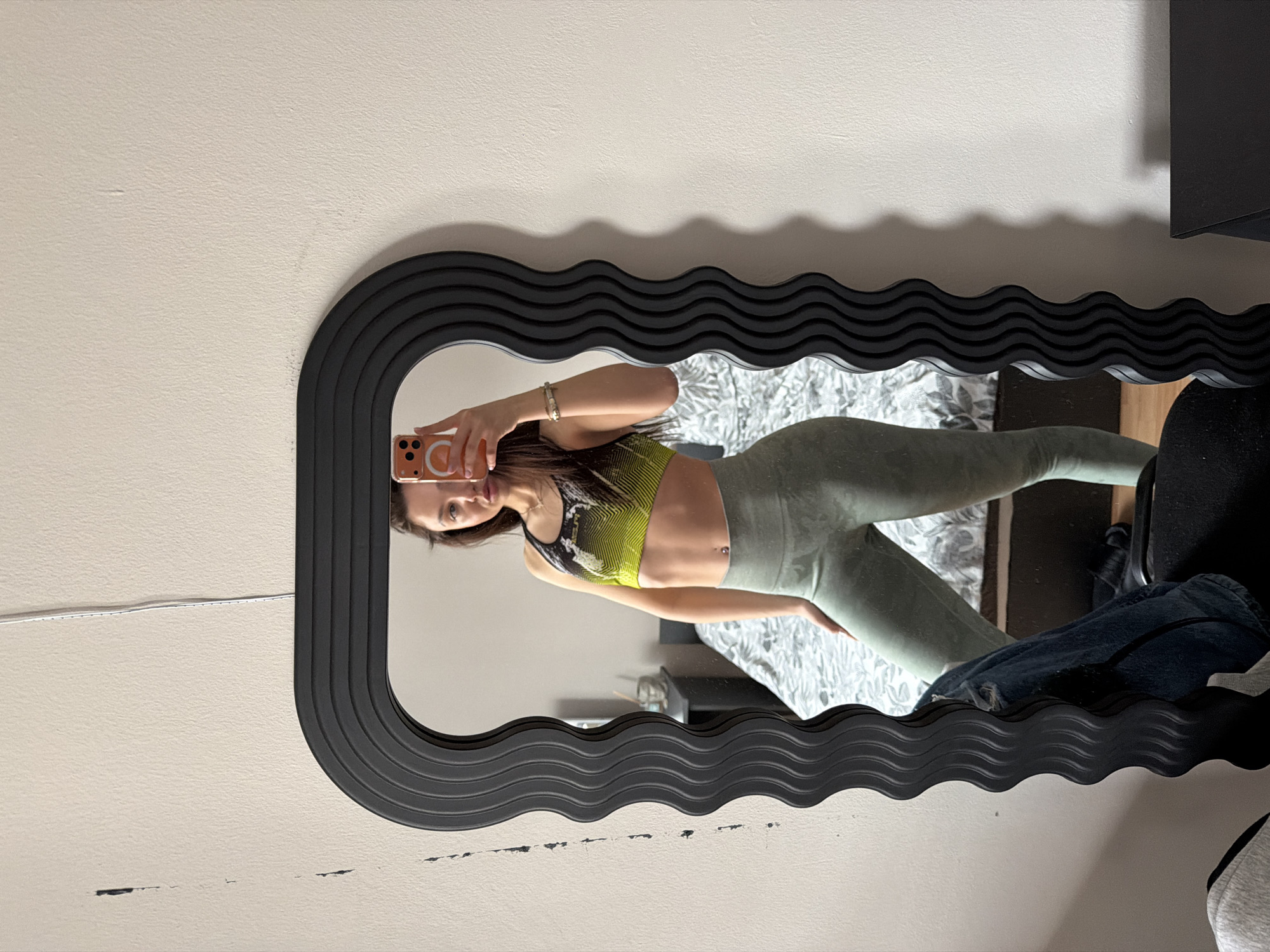 A young woman is taking a selfie in a mirror. She is wearing a black and yellow sports bra and green leggings. The mirror has a black, wavy frame and is hanging on a light beige wall. A white cable is visible on the upper left of the mirror. The woman is holding an orange phone and looking at the camera. The location is indoors. The background features a bed with a white and grey patterned comforter and a black desk and chair. There is no evidence to suggest that the photo was taken in Sofia, Bulgaria.