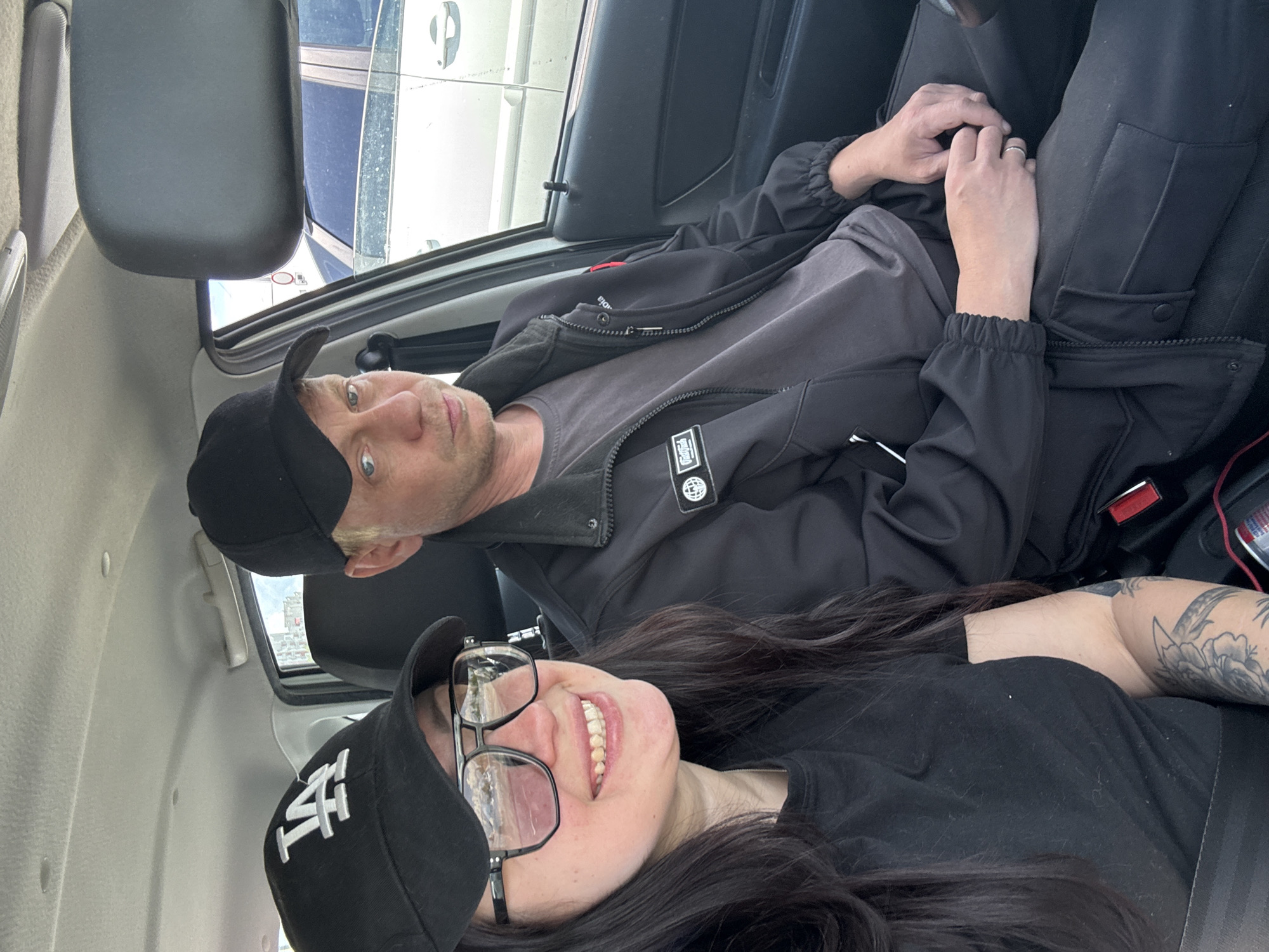 Two people, a man and a woman, are in the front seats of a vehicle. The man, wearing a black baseball cap, a gray t-shirt, and a black jacket, is sitting upright. His hands are clasped in his lap. The woman, wearing a black baseball cap with the letters "LA" on it and glasses, is sitting in the passenger seat. She is smiling. The vehicle appears to be parked, and the location is Unknown, Unknown. The image is slightly rotated.