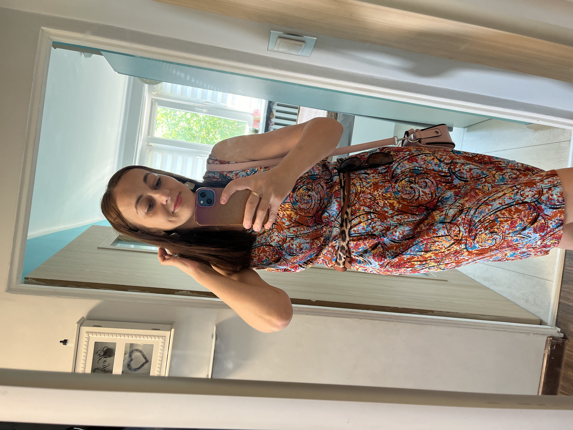 A woman is taking a mirror selfie in an indoor setting. She is wearing a colorful, sleeveless dress with a swirling pattern, a leopard print belt, and a pink crossbody bag. She is holding a smartphone in her right hand and touching her hair with her left. The mirror reflects a room with a window, blinds, and trees visible outside. There is also a white wall-mounted cabinet with decorative elements in the reflection.