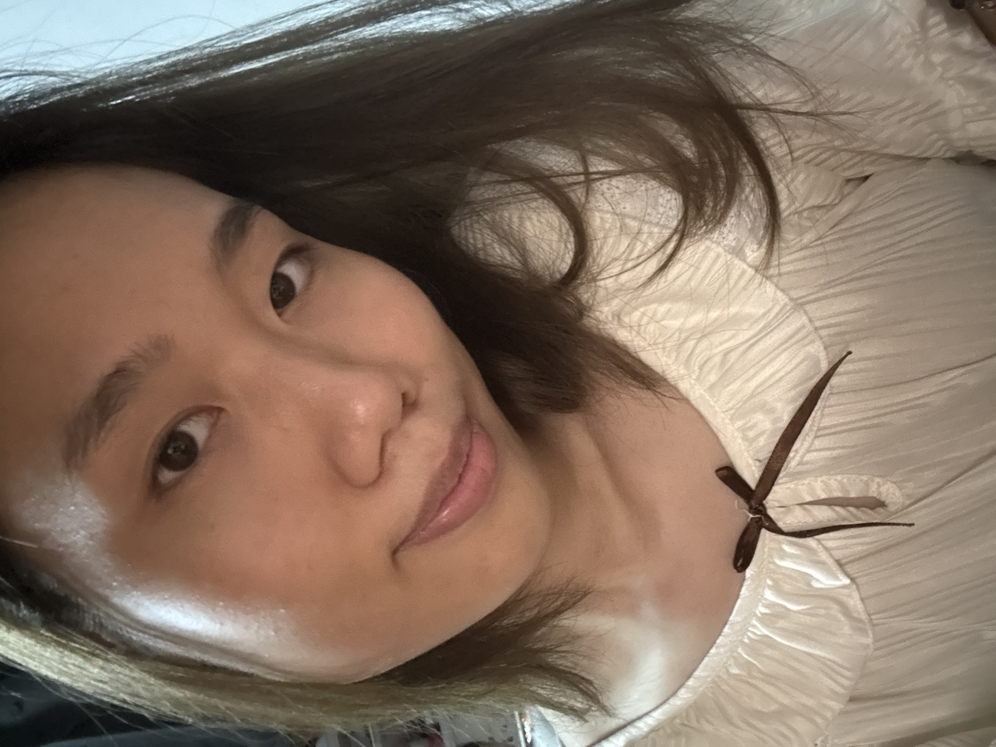 A close-up, selfie-style image of a woman with brown hair, fair skin, and Asian features. She is wearing a white, textured blouse with a brown ribbon tied at the collar. The background is out of focus and indistinct, providing no clear indication of location. There is no discernible information to place this image in the city of Nan'an District, China.