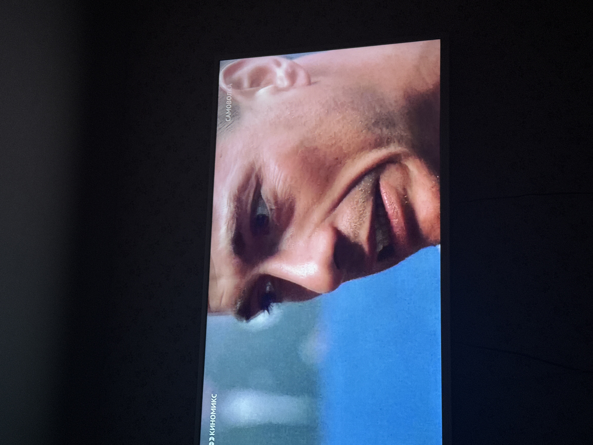 The image shows a close-up of a television screen displaying a distorted, upside-down image of a man's face. Text in Russian "Киномикс" (Kinomix) is vertically oriented and visible on the left edge of the screen, and "САМОВОЛКА" (SAMOVOLKA) is visible on the top. The screen is surrounded by a dark, possibly black, background, suggesting the image was taken in a dimly lit environment. No specific location is discernible from the image, beyond the fact that it could be Russia based on the Cyrillic text. Mytishchi is not determinable from the image.