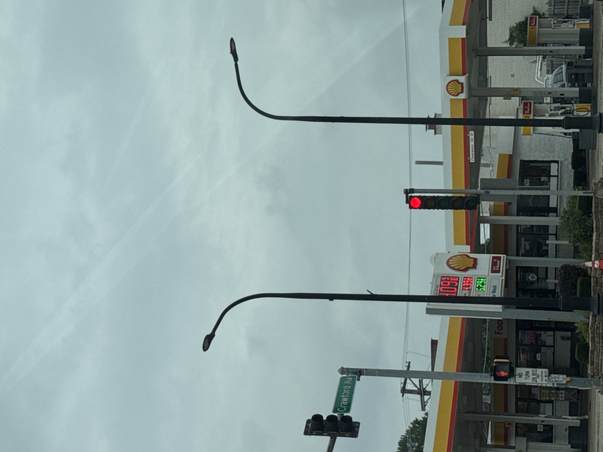 The image depicts a street scene at a Shell gas station in Lincolnwood, Illinois, United States of America. The sky is overcast with visible clouds. Two streetlights with curved poles stand prominently. A traffic light is visible on the left, displaying a red signal. A street sign indicates "Crawford Av." to the left of the traffic light. The gas station features the Shell logo and a gas price display showing prices of 4.09, 4.39, and 5.29. Fuel pumps are visible under the covered area of the station. A "No Turn on Red" sign for pedestrians is also present. The overall environment appears to be a typical urban streetscape with commercial buildings.