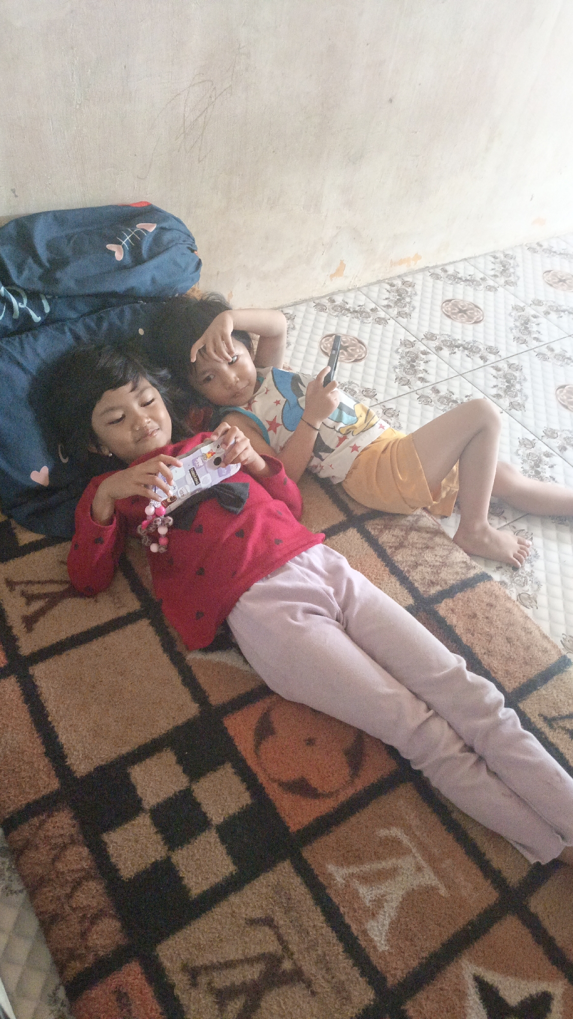 Two young girls are lying on a patterned rug in a room. The girl in the foreground on the left is wearing a red long-sleeved shirt with black polka dots and light purple pants. She is holding a white object with cartoon characters on it, possibly a toy or a phone case, and a string of pink beads. Her eyes are partially closed. The girl behind her on the right is wearing a white and blue tank top with red stars and yellow shorts. She is holding a black smartphone in her right hand and looking towards the viewer. Her left arm is bent at the elbow with her hand near her face. They are lying on a rug with a geometric pattern in brown, black, and orange. Behind the rug, there is a tiled floor with a floral pattern. To the left of the girls, there is a dark blue pillow with a white fish skeleton design. The wall behind them is light-colored and appears to be stucco. This scene is likely in a residential setting.