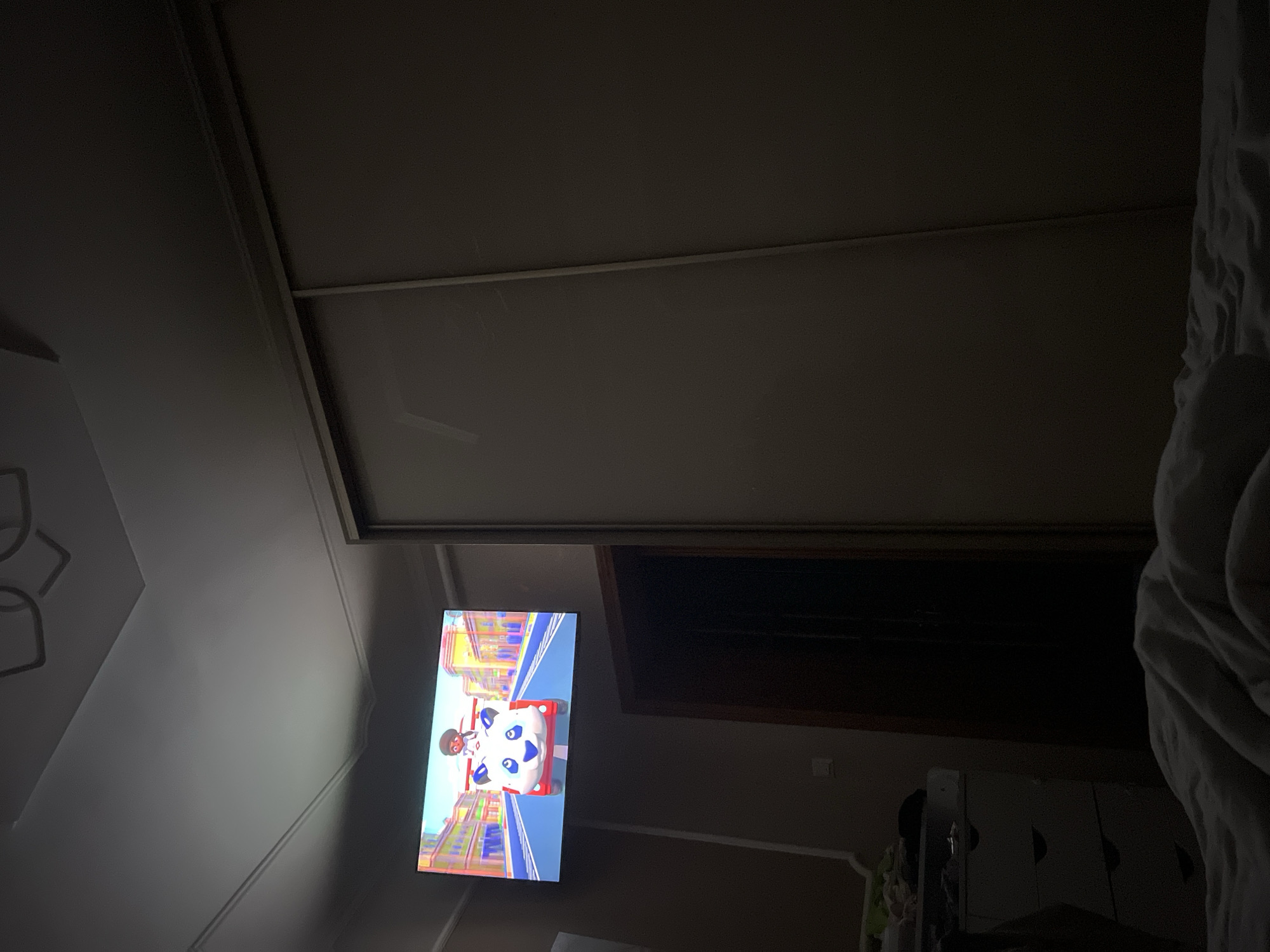 In a dimly lit room, a television screen displays a colorful cartoon with a character driving a panda-shaped vehicle. The TV is mounted on the wall above a dresser with several drawers, which is partially visible on the left side of the frame. To the right, a large wardrobe with sliding doors dominates the space. A doorway, appearing to lead to another room, is visible between the TV and the wardrobe. The floor is covered by a rumpled white blanket, suggesting a bedroom setting. The ceiling has decorative molding and a light fixture. The image does not provide any specific information about the location being Vanchykivtsi, Ukraine.