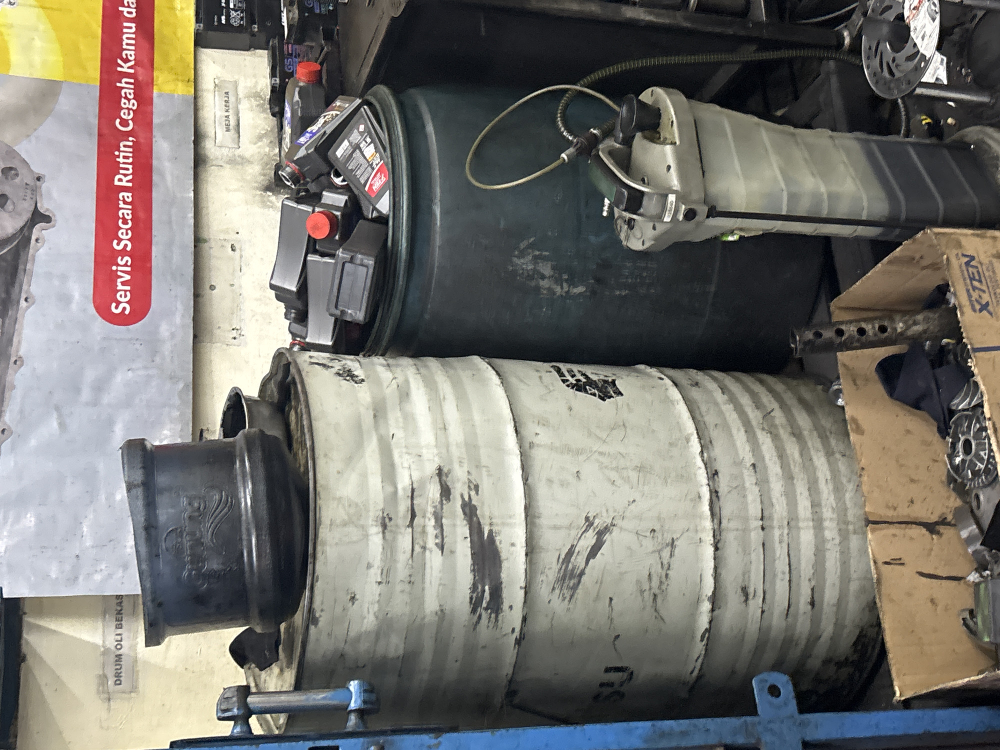 The image shows a cluttered workshop environment, likely in North Jakarta, Indonesia, given the text on the sign that reads "Servis Secara Rutin, Cegah Kamu..." which translates to "Regular Service, Prevent You...". In the foreground, a large, grey fluid drainer or extractor is visible, with its transparent tank filled with dark liquid. To the left, two stacked, large, weathered metal drums are prominent. The upper drum has a black metal lid or funnel-like object on top, with some text visible on it, possibly a brand name. The lower drum has some white markings and appears to be stained. Behind the drums, a large dark green plastic barrel is filled with several black plastic bottles, presumably oil or lubricant containers, with red caps. A few of these bottles have visible labels. Above, a large banner with red text on a white background is partially visible, with the message "Servis Secara Rutin, Cegah Kamu da...". To the far left, a blue metal support structure is visible. A cardboard box containing metal parts and tools is in the lower left corner of the image. The overall impression is of a busy, functional automotive workshop.