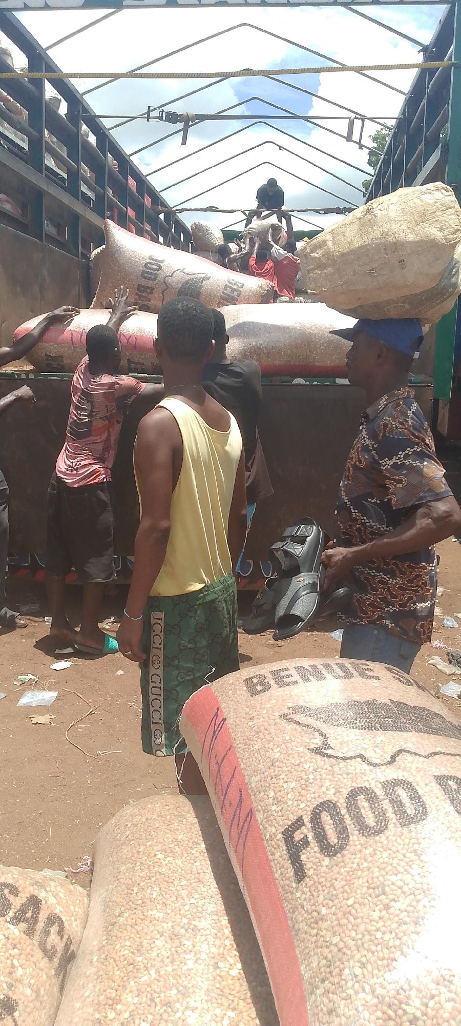 The image depicts a busy outdoor scene in Tor-Donga, Nigeria, likely a market or loading area. In the foreground, several large burlap sacks filled with grain are visible, some bearing markings like "BENUE" and "FOOD". A man wearing a blue cap and a patterned shirt is seen with a sack balanced on his head, while another man in a yellow tank top and patterned shorts stands nearby. Behind them, a truck is being loaded with more sacks, with multiple individuals engaged in the activity. Some people are carrying sacks, while others appear to be organizing them. Sandals and other footwear are scattered on the ground. The environment is dusty and appears to be an open-air space. The overall impression is one of active commerce and labor.
