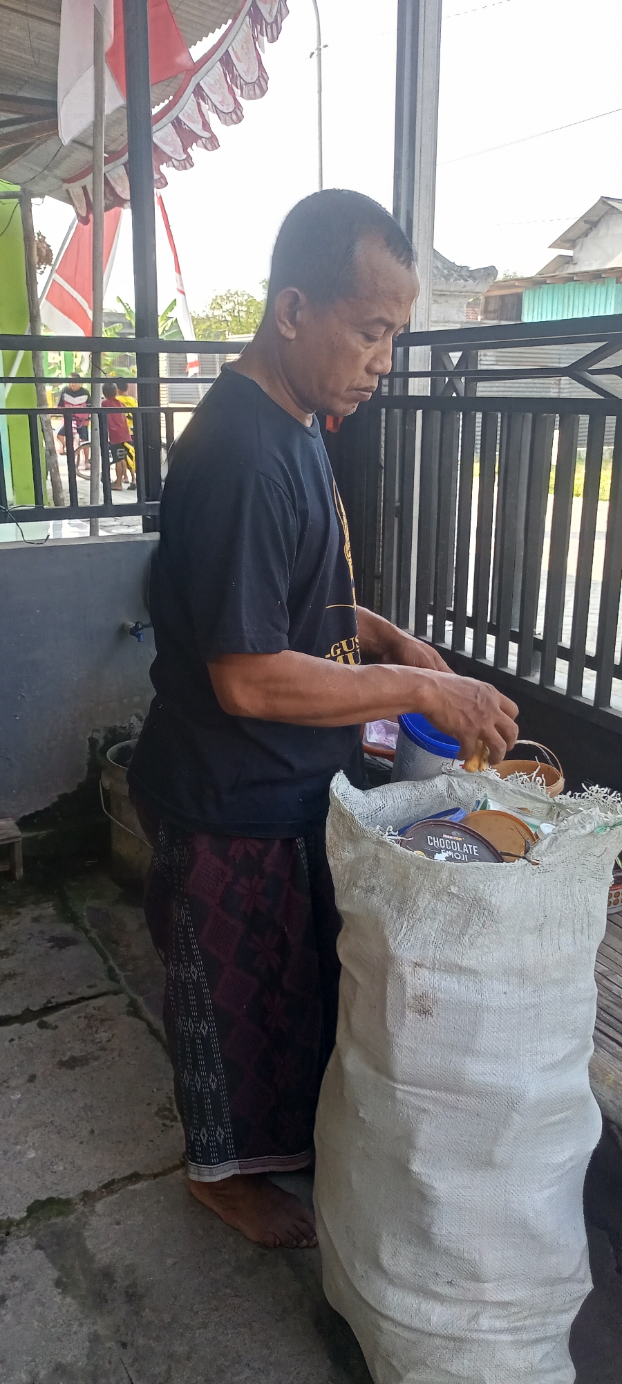 In Gempolklutuk, Indonesia, a man stands at a concrete ground level outdoors, near a fence. He is wearing a dark t-shirt and a patterned sarong. The man is holding and sorting through the contents of a large, off-white woven sack. Inside the sack are various cylindrical containers, some with visible labels like "CHOCOLATE". He is focused on this task, with his hands actively engaged with the items. To his left, a blue bucket and a metal barrel are visible, suggesting a utilitarian or work-related environment. Behind him, a black metal fence with vertical bars separates the immediate area from the background. The background includes elements of buildings with corrugated metal roofs and red and white festive decorations strung overhead, possibly indicating a local celebration. Children can be seen in the distance through the fence. The overall scene depicts a man engaged in sorting materials from a large sack in an outdoor, possibly semi-public space in Indonesia.