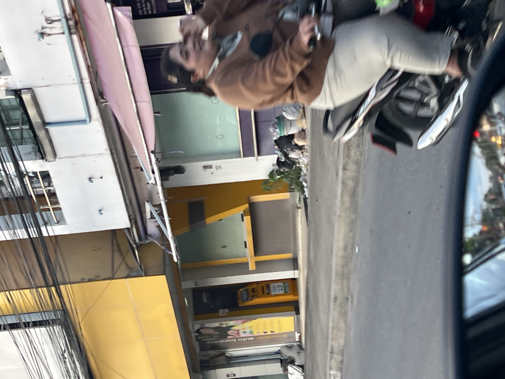 The scene depicts a person riding a motorcycle in Baan Bo Phut, Thailand. The individual is wearing a brown jacket and light-colored pants. The motorcycle is moving along a road adjacent to a building with yellow and white facades, possibly a commercial establishment with an ATM or banking facility. Cables are visible overhead, and there is debris or waste near the building. The angle suggests the photo was taken from a vehicle's side mirror.