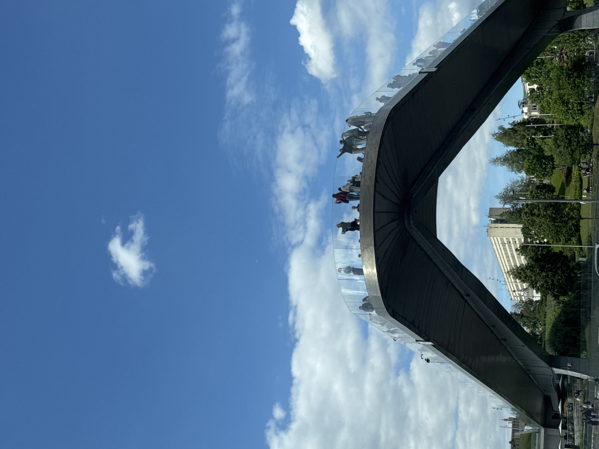 The image shows the Zaryadye Park bridge in Moscow, Russia, seen from an upward angle. The bridge's unusual, boomerang-like design is filled with people strolling along its path. Clear safety glass lines the bridge's edges. The sky behind the bridge is partly cloudy. Trees and buildings are visible behind the bridge, indicating an urban park environment.