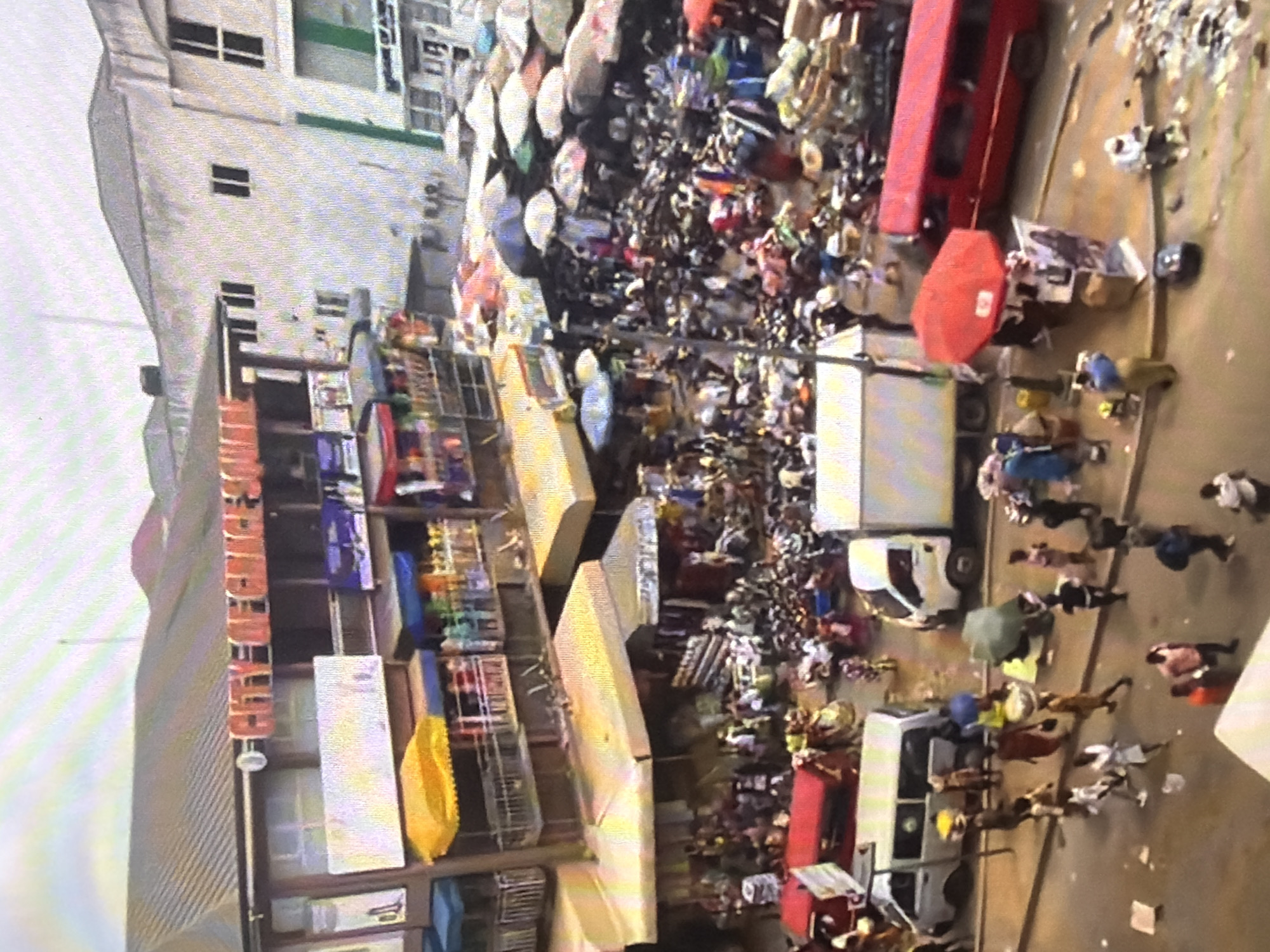 The image depicts a bustling market scene in Bukuru, Nigeria, with a crowd of people engaged in commerce. A multi-story building with shops occupies the left side of the frame, featuring awnings and displays of goods. The street level is filled with vendors and shoppers, with various vehicles, including vans and buses, present. Umbrellas are used to provide shade over stalls in the crowded market area. The environment appears to be an outdoor market with a mix of commercial and residential structures.
