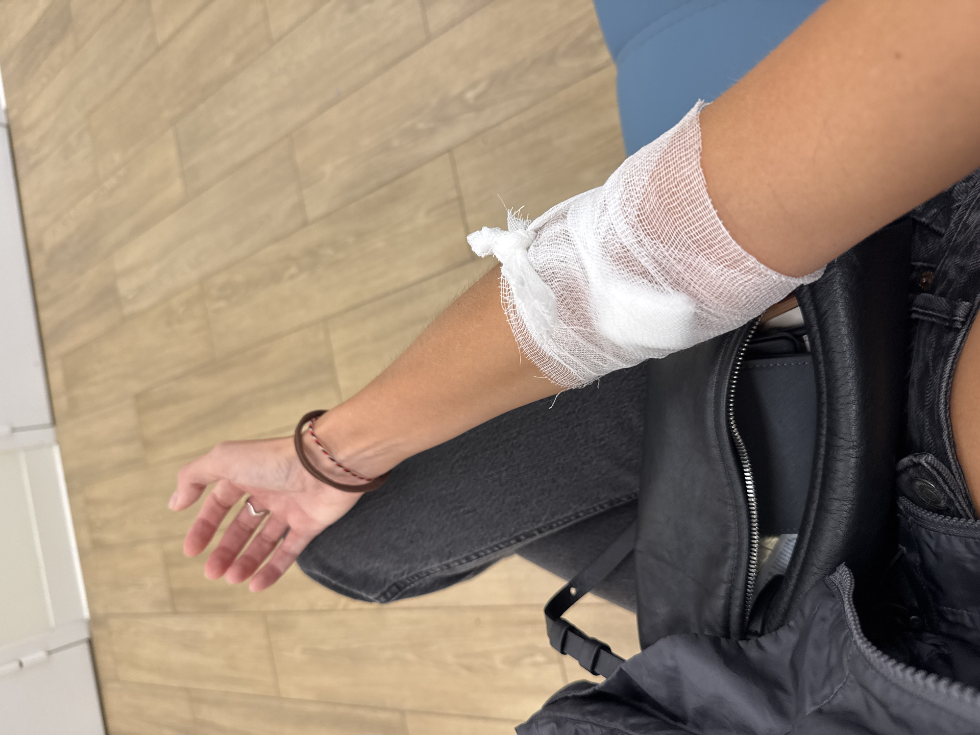 A person's left arm is bandaged at the wrist, resting on their lap. The person is wearing dark denim jeans and a dark jacket. A black leather handbag is placed on the person's lap. The background consists of a light-colored wooden floor and a light-colored wall. The image is taken from a first-person perspective, looking down at the person's arm and lap. The image does not depict any specific location in Moscow, Russia.