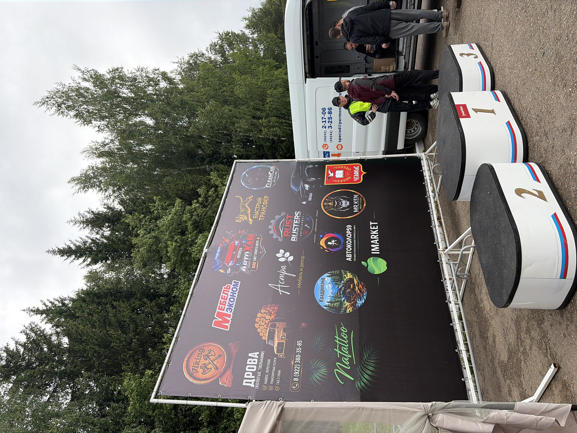 This image depicts an outdoor scene in Прикамский, Russia, featuring a large sponsor banner, three podiums, and a white commercial van, with a group of six men gathered nearby. The central subject is a large, rectangular, black banner supported by a white metal frame. It displays numerous logos and text in various colors, indicative of sponsors or participants. Notable elements on the banner include: a "Viking" logo with a wooden barrel and crossed axes; "МЕБЕЛЬ ЭКОНОМ" (Furniture Economy) text; "АВТОХАМ" (Avto Kham) with a red car and license plate X560AX 91; a tiger logo; "БЫСТРЫЙ ТРАНСФЕР" (Fast Transfer); "CLEANCAR DETAILING"; "ДРОВА" (Firewood) with a phone number (8 (922) 360-35-85) and an illustration of a log truck; "Астра" (Astra) for furniture and decor; "RUST BUSTERS АВТОСЕРВИС" (Rust Busters Auto Service); "Natalloo" in green script with palm leaves; "Квадролэнд" (Quadroland) with an ATV graphic; "АВТОКОЛОР59" (AutoColor59) for professional car painting; "MR. KEN PRINT STUDIO" with a crowned gorilla head; "ЧГАФКИС" (CHGAFKIS); and "IMARKET" with a green apple logo. In front of the banner, three white, oval-shaped podium blocks are arranged sequentially from right to left, numbered "1", "2", and "3" in gold/brown font on their fronts. Each podium has a black textured top surface and a horizontal stripe in the colors of the Russian flag (white, blue, red) across its lower front. The "Альфа Банк" (Alfa Bank) logo is visible on podium number 1. To the right of the banner, a white commercial van is parked, with its rear passenger-side door open, revealing boxes or items inside. The van's side displays two phone numbers ((34241) 2-17-06 and (34241) 3-25-86) and a partially visible email address (specod@permon.ru). Six adult males are present in the scene. Three men are standing near the open van door: one in a black jacket with a yellow reflective vest, one in a dark red hoodie with hands behind his back, and one in a dark hoodie and gray sweatpants. Two other men are seated inside the van, one of whom is looking at a smartphone. A sixth man is partially visible further to the back of the van. All individuals are casually dressed. The ground is composed of light brown, compacted dirt or gravel. On the far left, a portion of a light-colored tent or canopy structure with sheer fabric is visible. The background consists of a dense forest with a mix of tall coniferous and deciduous trees,