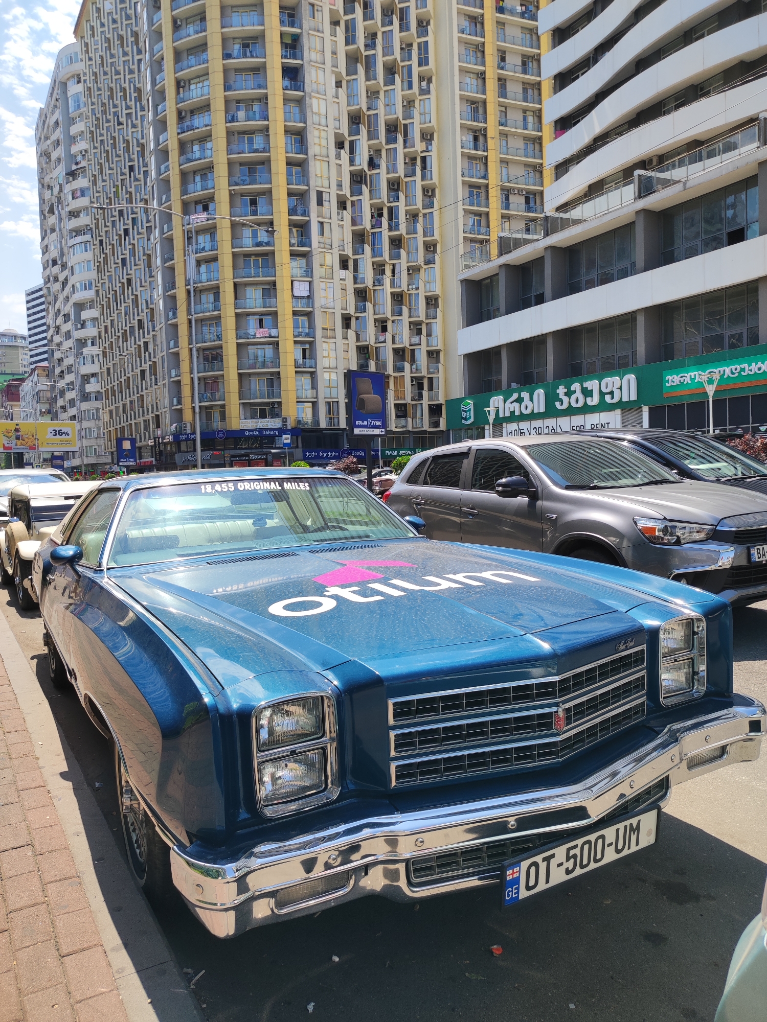 In Batumi, Georgia, a blue Chevrolet Monte Carlo is parked on a street, facing the viewer. The car has a chrome bumper and grille, and a pink graphic with white text is visible on the hood. A license plate with "GE OT-500-UM" is mounted on the front bumper. Behind the Monte Carlo, a gray SUV and another car are parked. Tall buildings with many windows line the street, and there are signs with Georgian text on them. The sky is cloudy.