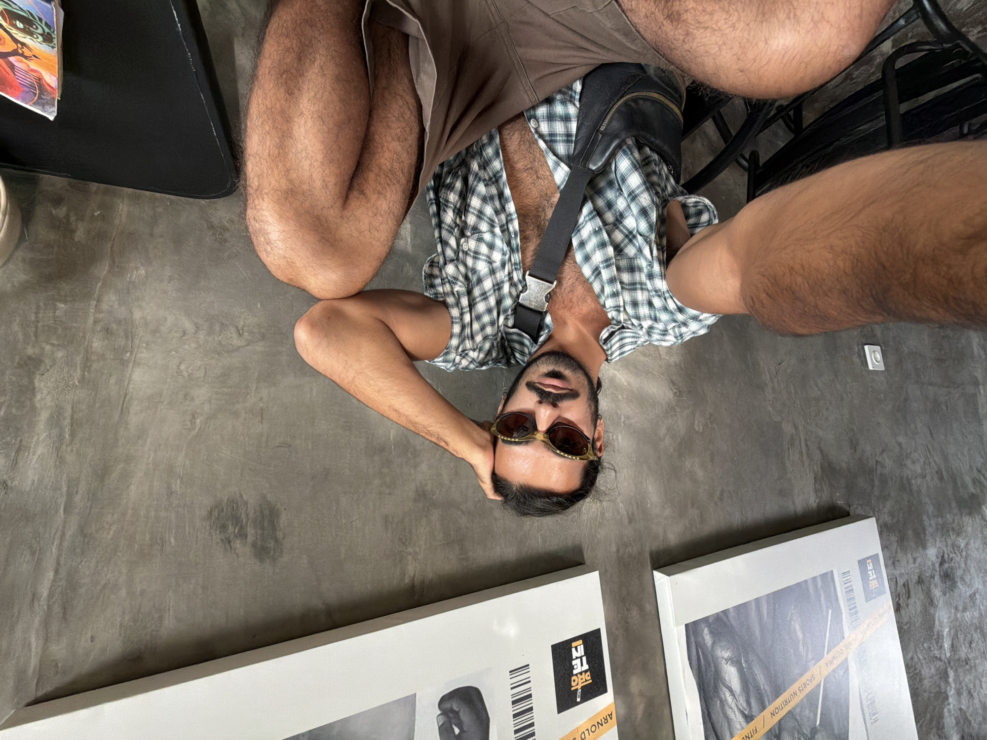 The image depicts a man sitting indoors, in what appears to be a cafe or restaurant with a concrete wall as the backdrop. The man is wearing a green and white plaid short-sleeved shirt, unbuttoned to reveal his chest, and brown shorts. He has sunglasses on and his hand is resting on his head. A black sling bag is worn across his chest. In the background, there are two framed posters on the wall, one of which features text related to "SPORTS NUTRITION" and "ARNOLD". The lighting suggests an indoor environment, possibly with ambient light. The overall scene is casual and relaxed. The image is taken from a slightly low angle, looking up at the man.
