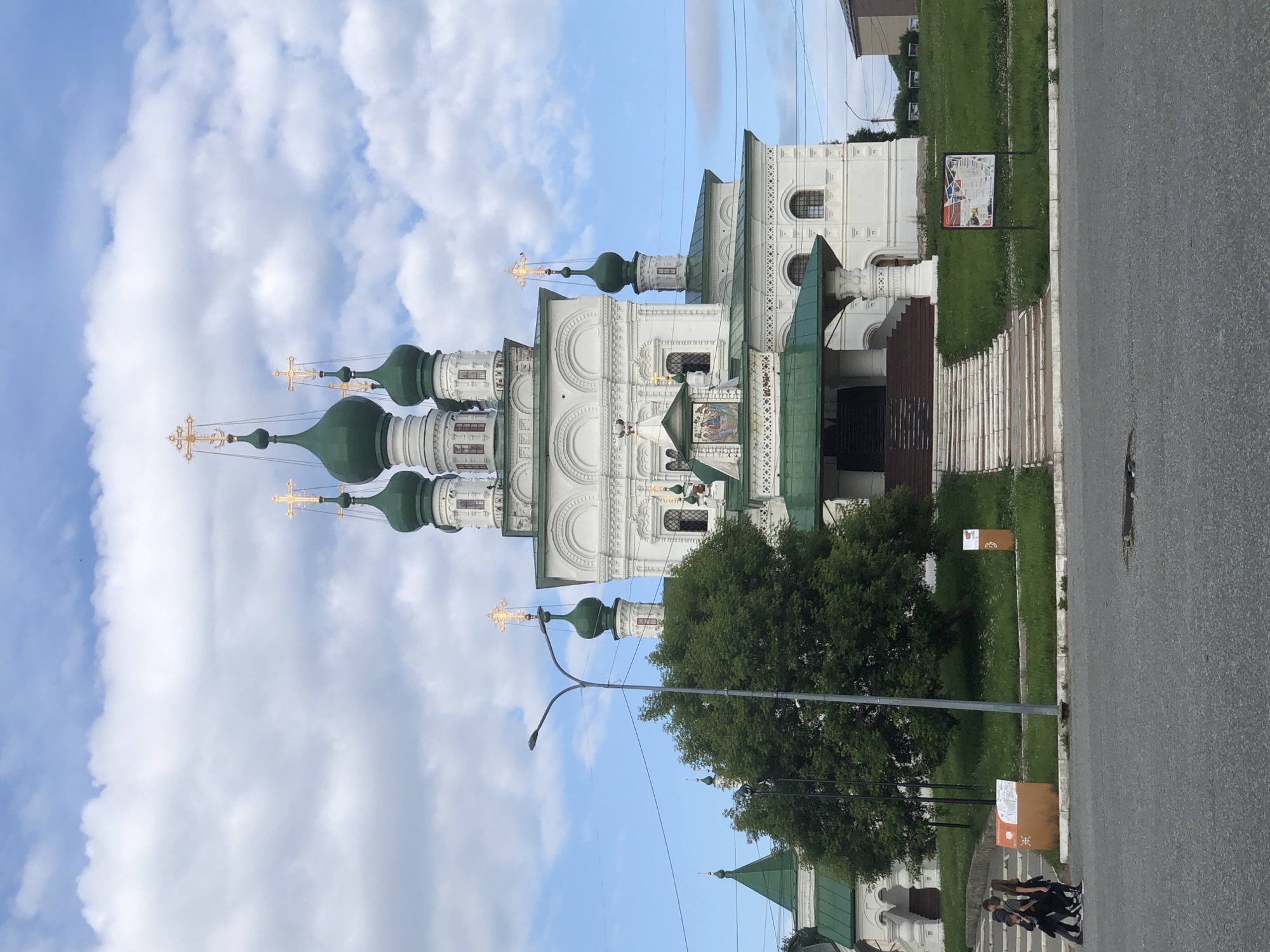 This media file, oriented vertically, captures a view of a white orthodox church with green domes topped by golden crosses. The church features intricate architectural details and is partially obscured by a large, lush green tree. In the foreground, a paved road runs alongside a grassy area where several signs are posted. Some of these signs appear to have informational graphics or text, though they are not clearly legible. A few people are walking on the sidewalk near the road and the church entrance. The sky is blue with scattered white clouds, suggesting fair weather, possibly in the late afternoon or early evening due to the soft lighting. The overall scene depicts a tranquil setting in Solikamsk, Russia, showcasing religious architecture and a public space.