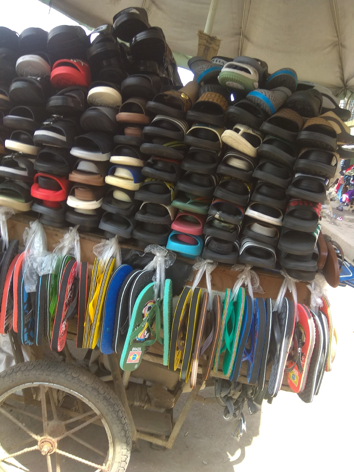 This is a medium-shot, eye-level, full-color photograph of a street vendor's cart in Jalingo, Nigeria, overflowing with a variety of footwear, primarily flip-flops and sandals. The cart itself is a rustic wooden structure with a large, weathered spoked wheel visible on the left side. Above the cart, a light-colored tarp or awning provides shade.
The footwear is meticulously organized and displayed in stacked layers on shelves, and also hanging in rows from hooks. The stacks of sandals and flip-flops are predominantly black, with some white, red, and brown accents. Colors are more vibrant in the hanging section, featuring yellow, blue, green, red, and patterned straps. One pair of green flip-flops prominently displays a Brazilian flag and a soccer ball design. The footwear appears new and in good condition.
The ground beneath the cart is composed of dirt or sand. The overall impression is one of a bustling market or roadside stall, offering a wide selection of affordable footwear. No people are clearly visible in the immediate foreground or on the cart, though the context suggests a vendor is present. The lighting suggests it is daytime, likely mid-morning or afternoon.
