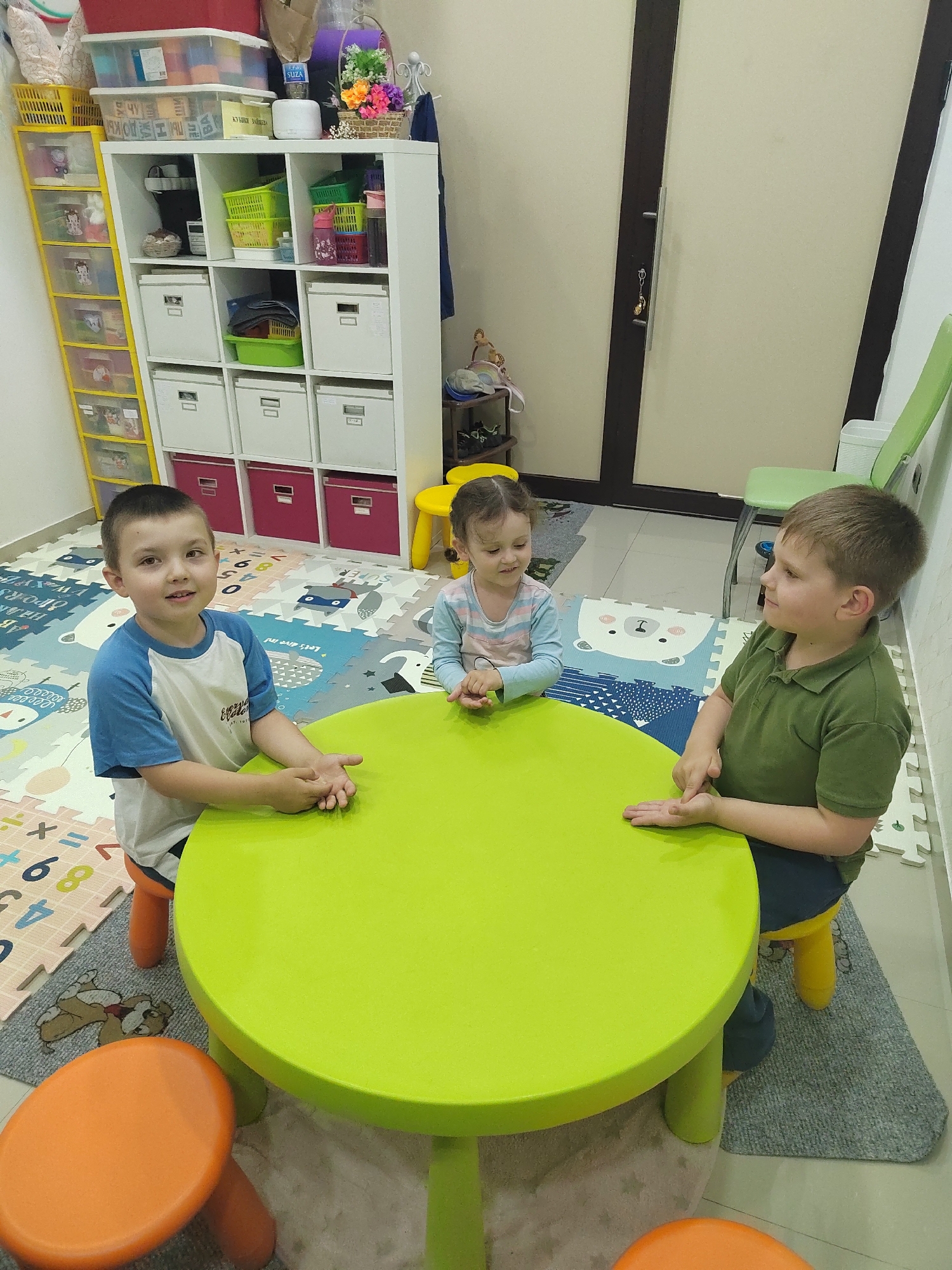 Three young children sit at a round, green table. A boy in a blue and white shirt faces the camera, resting his hands on the table. To his right, a girl with pigtails sits with her hands clasped together. On the other side of the table, another boy in a green polo shirt looks towards the girl.
The setting appears to be a playroom or classroom. The floor is covered with interlocking foam mats with letters and numbers. Behind the children, there is a white storage unit filled with colorful bins and boxes. A yellow plastic drawer unit is visible on the left. A closed door with a vertical dark accent is on the right. A green chair is partially visible against the wall. A small yellow stool sits near the storage unit.
The image does not contain information to determine the location in Podgorica, Montenegro.