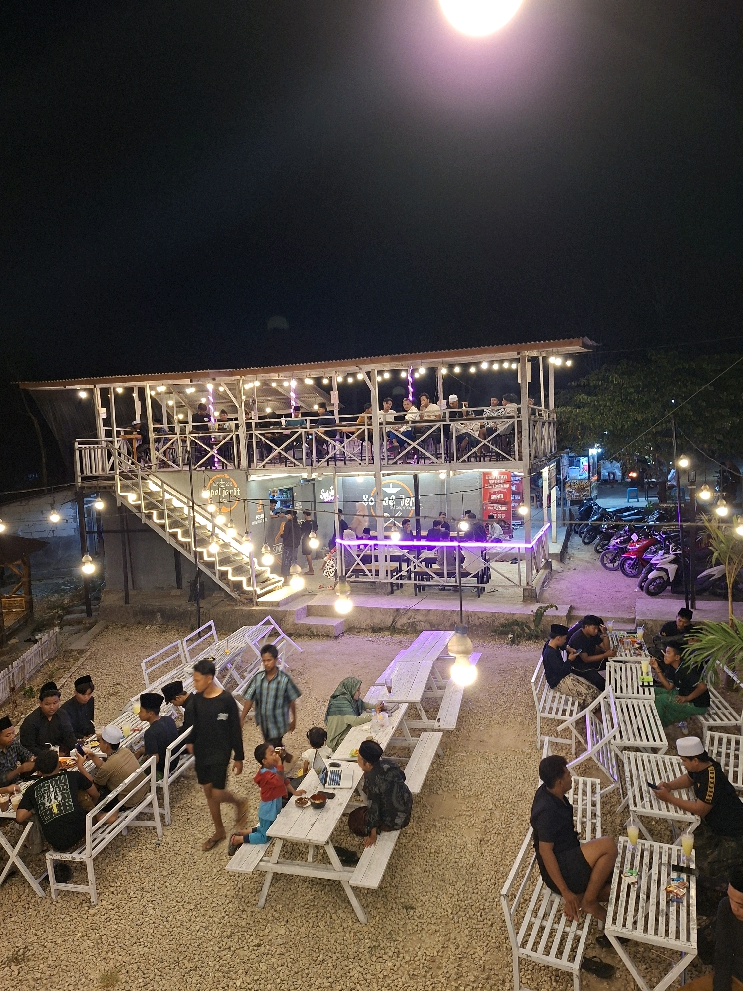 This is an aerial, night-time photograph of an outdoor dining establishment in Lantek Barat, Indonesia. The establishment features a two-story structure with a rooftop dining area, accessed by a brightly lit staircase. The grounds are covered in gravel, and numerous picnic tables with white benches are set up for patrons.
The lower level has additional seating areas, and a food stall or counter is visible. The upper level is enclosed with railings and also contains dining tables and seating. The entire area is illuminated by strings of hanging light bulbs, creating a warm and inviting atmosphere.
Several groups of people are seen dining and socializing. On the lower level, a group of men and boys are seated at picnic tables, some engaged in conversation, while others appear to be eating. A child in a red shirt and blue pants is walking near a table. On the upper level, more patrons are seated at tables. In the background, a row of motorcycles is parked.
The scene depicts a busy evening at a local eatery, with people enjoying meals and the ambiance provided by the lighting and outdoor setting.
