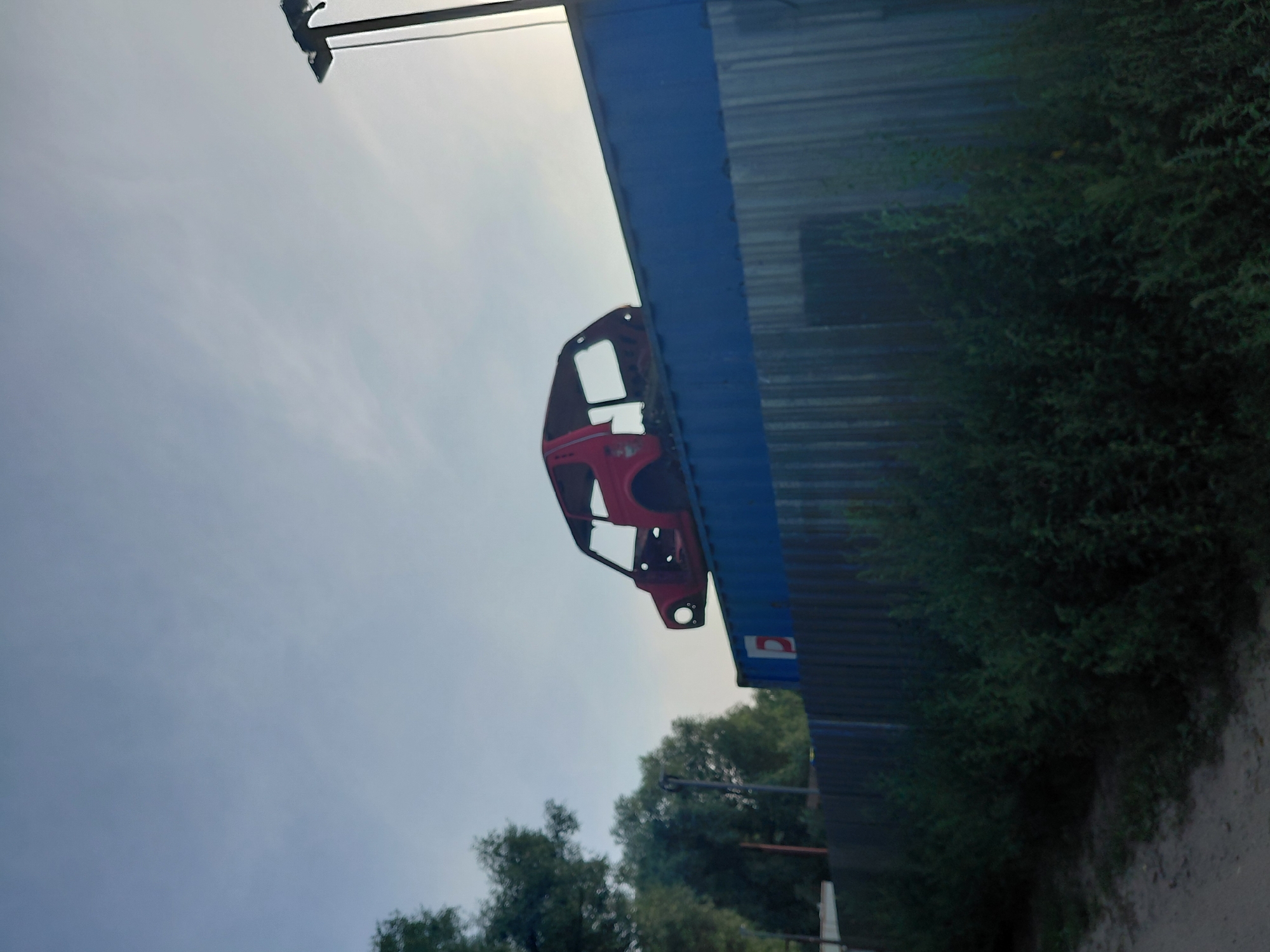 A bright red, stripped-down car body sits on top of a blue shipping container. The car has no windows, doors, or wheels. It is positioned facing left with its front end slightly elevated. The shipping container is resting on top of a corrugated metal fence, which extends across the lower portion of the image. To the left of the fence, dense green foliage is visible. In the background, trees and a light pole can be seen under a pale sky. The scene is likely outdoors, possibly in an industrial area or a junkyard.