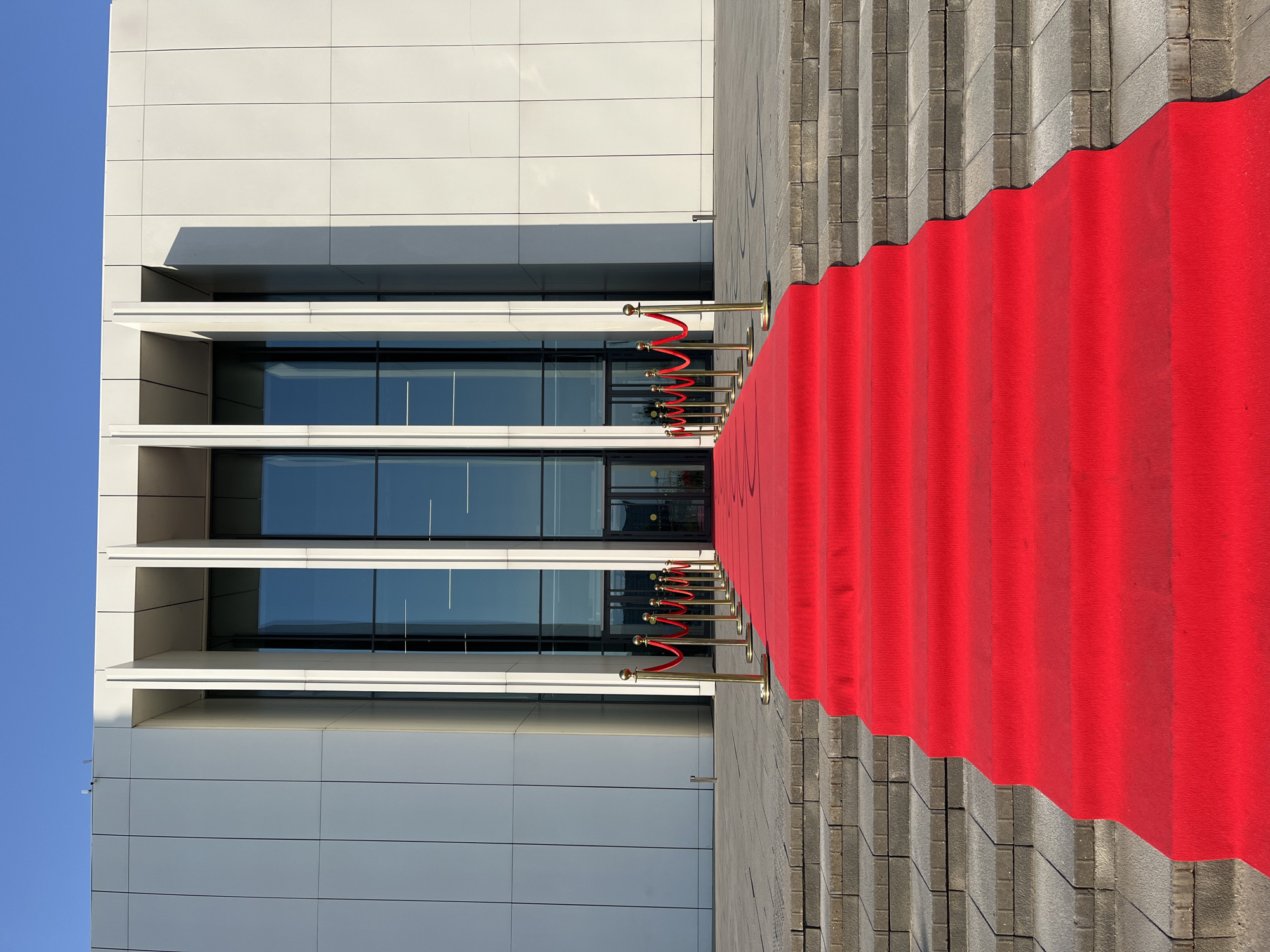 The image depicts a grand entrance to a modern building, likely a cultural or event venue, situated in Nizhny Novgorod, Russia. A vibrant red carpet, unfurled across a set of wide, grey stone stairs, leads towards the building's entrance. The carpet is bordered by polished gold stanchions and red velvet ropes, creating a sense of occasion and exclusivity. The building itself is characterized by its sleek, contemporary architecture, featuring a facade of white panels and tall, narrow glass windows that reflect the clear blue sky. The overall scene is one of elegance and anticipation, suggesting a formal event or VIP reception. The setting appears to be outdoors, under bright daylight, with the architecture and the clean lines of the red carpet signifying a meticulously prepared space.