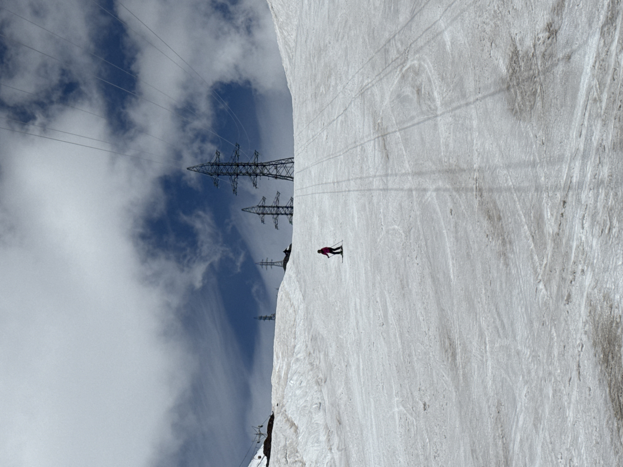 This image, taken in Terskol, Russia, captures a skier descending a snow-covered slope against a backdrop of a bright blue sky with wispy white clouds. The skier, dressed in a pink jacket and black pants, is positioned on the right side of the frame, mid-descent. Their skis are parallel, and they appear to be in a controlled turn.
Dominating the left side of the image are towering electricity pylons with multiple lines stretching across the sky. These structures suggest the proximity of infrastructure to this natural, mountainous landscape. The snow on the slope shows visible tracks, indicating recent activity and the presence of other skiers or snowboarders. The sunlight casts shadows, suggesting it is daytime, likely mid-morning to afternoon, given the brightness of the sky and the definition of the shadows. The overall scene conveys a sense of outdoor recreation and the vastness of the mountainous terrain. There is no visible text in the image.