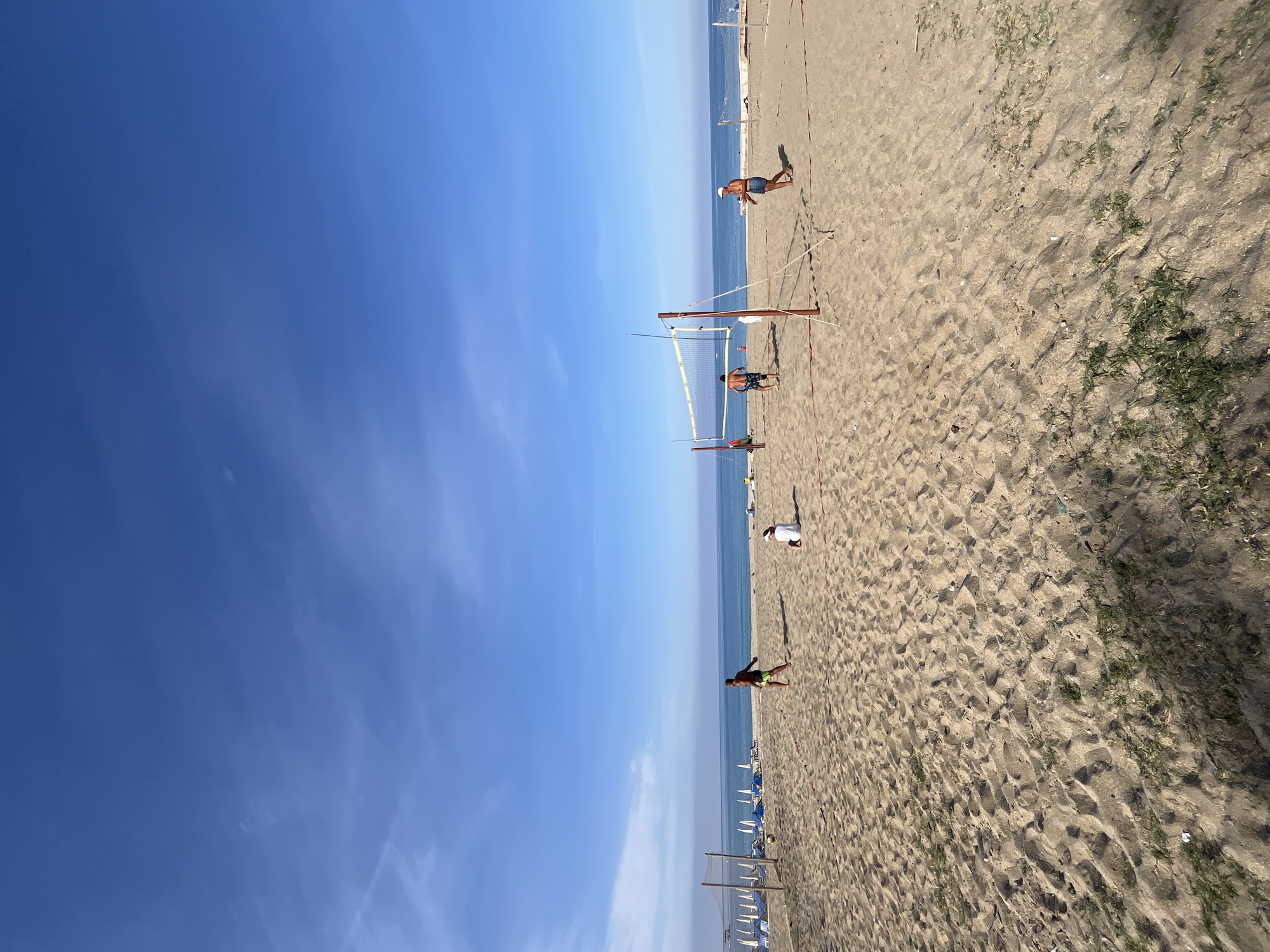 The image shows a beach scene in Alanya, Turkey. The sky is clear blue. People are playing beach volleyball near a net in the sand. There are beach umbrellas and chairs set up along the shore. The sand is light brown and textured. Some scattered vegetation is visible on the sand.