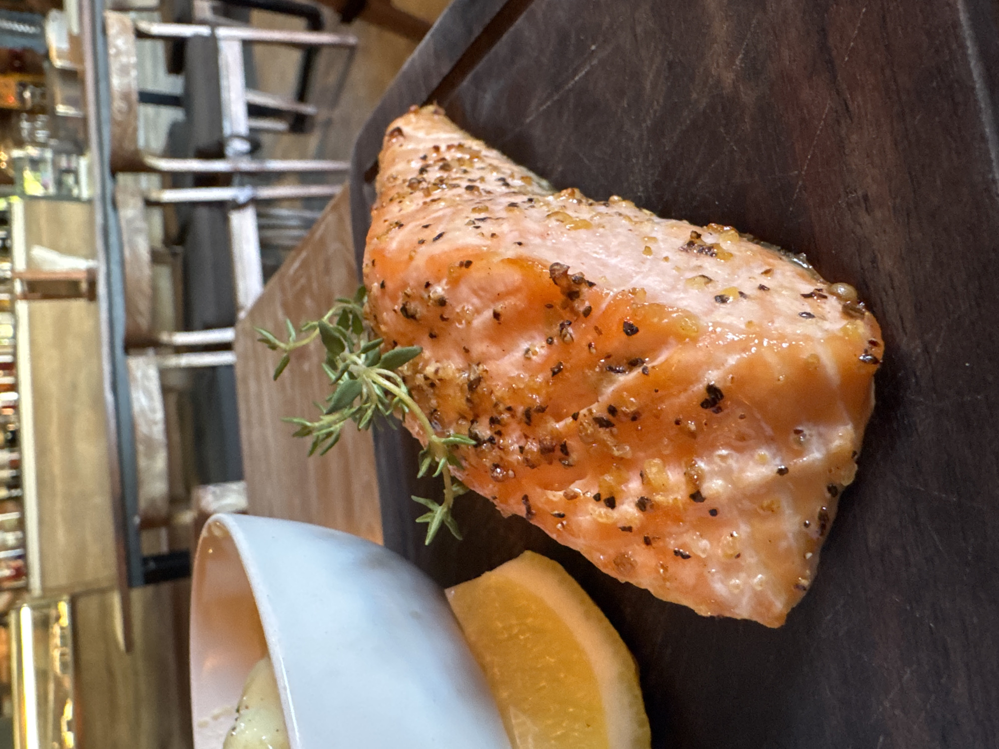 A salmon fillet, garnished with a sprig of thyme and a lemon wedge, rests on a dark, possibly wooden, serving board. A white, angled dish or bowl is partially visible near the lemon. The fillet has a browned, seasoned surface with visible peppercorns or other spices. The background suggests a restaurant setting with tables, chairs, and a display of bottles, possibly alcohol. The location in Kuta, Indonesia, is not explicitly visible in the image.