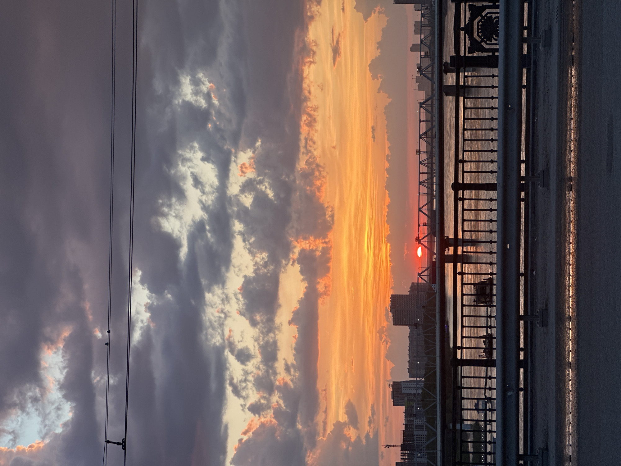 The image shows a sunset over the Ob River in Novosibirsk, Russia, as viewed from a bridge. The sun is partially obscured by buildings and the bridge's structure, casting a red glow on the water. The sky is filled with dramatic clouds, transitioning from dark gray on the left to orange and yellow near the sun. In the foreground, there is a metal railing and part of the bridge's pedestrian walkway. Power lines stretch across the left side of the frame, and there are birds perched on the powerlines. The buildings of Novosibirsk are visible in the distance.