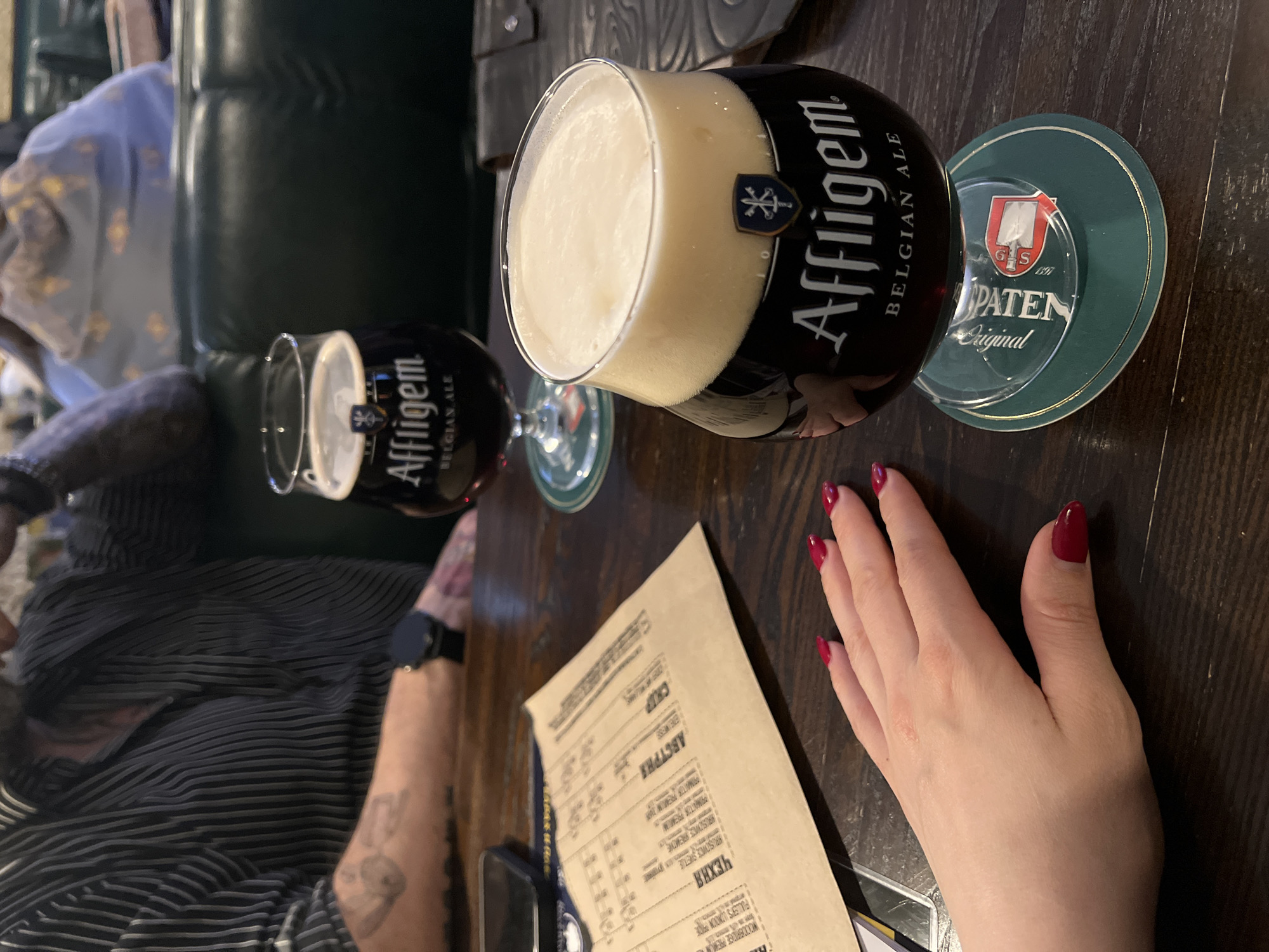 The image shows a table in what appears to be a pub or restaurant. Two glasses of Affligem Belgian Ale are visible. One glass is on a Spaten beer coaster. A menu with Russian text is on the table. A person's hand with red nail polish rests near the menu. Another person's arm with a watch and tattoos is also visible. The location cannot be determined from the image.