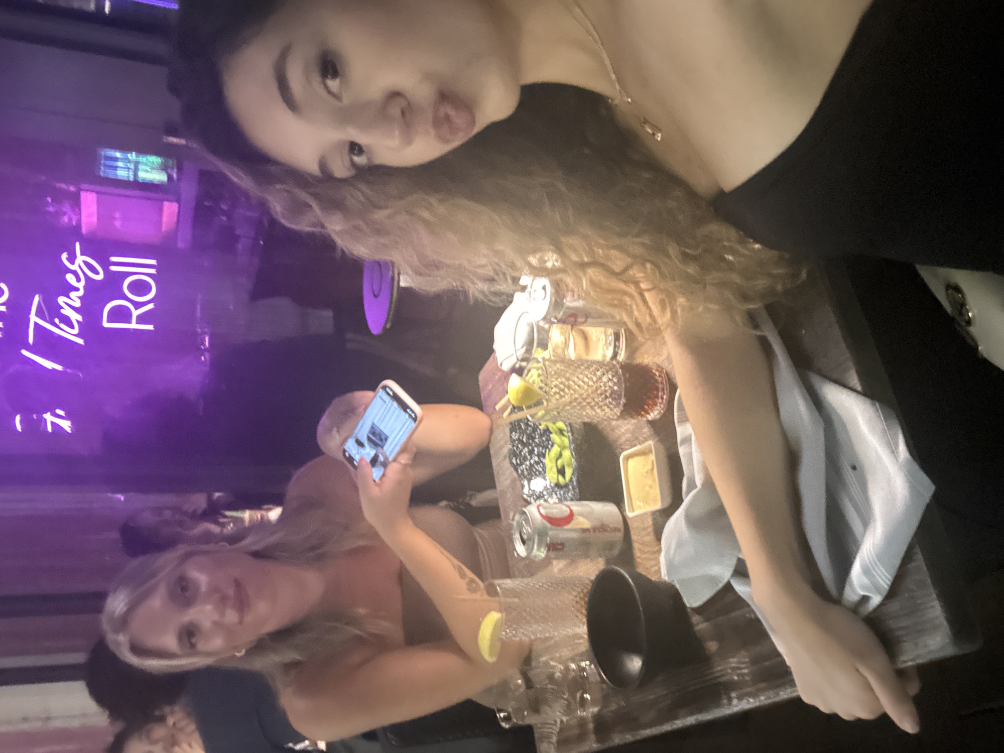 The image depicts two women sitting at a table in a dimly lit restaurant or bar, with a neon sign visible in the background displaying the words "Times Roll". The restaurant is located in Miami, Florida, United States of America. One woman is holding a smartphone and taking a photo, while the other woman is posing for the camera with pursed lips. On the table, there are several items, including a can, various glasses, a small dish with a yellow substance, and a folded light blue cloth. The table itself appears to be made of dark wood. The environment suggests a casual dining or nightlife setting.