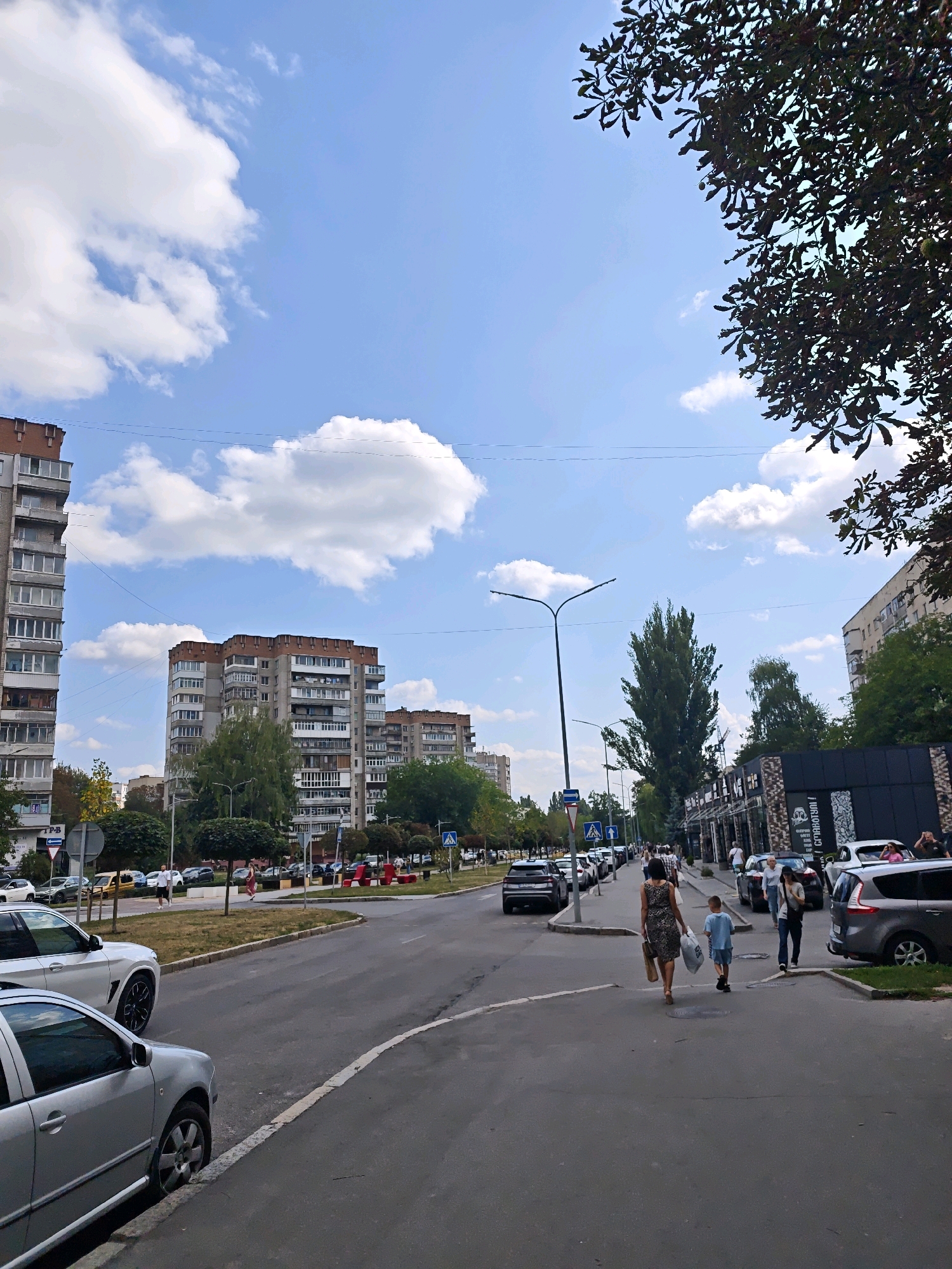 The image depicts a street scene in Vinnytsia, Ukraine, on a cloudy day. In the foreground, two cars are parked on the side of the road, one silver and one white. A paved road with a white line marking the lane curves to the right. Further down the street, several people are walking on the sidewalk, some carrying shopping bags. Buildings line the right side of the street, including what appears to be a commercial establishment with a dark facade and large windows. Across the street, there are tall apartment buildings. Trees and streetlights are also visible. The sky is blue with scattered white clouds.