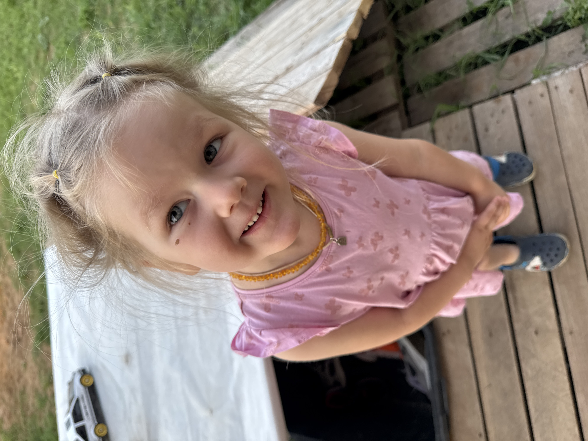 A young girl with blonde hair is the central subject of the photograph. Her hair is styled with small yellow rubber bands near the hairline. She has fair skin with a visible mole on her face, and she's smiling towards the camera. She is wearing a pink dress with a pattern of small shapes and a beaded necklace. She is standing on a wooden platform. There are blades of grass growing up between the slats of the platform. To the left of the girl is a white surface with a toy car, and behind her is a wooden structure. The photograph appears to be taken from a high angle, looking down at the subject. The scene appears to be outdoors. The location is Ферма, Russia.